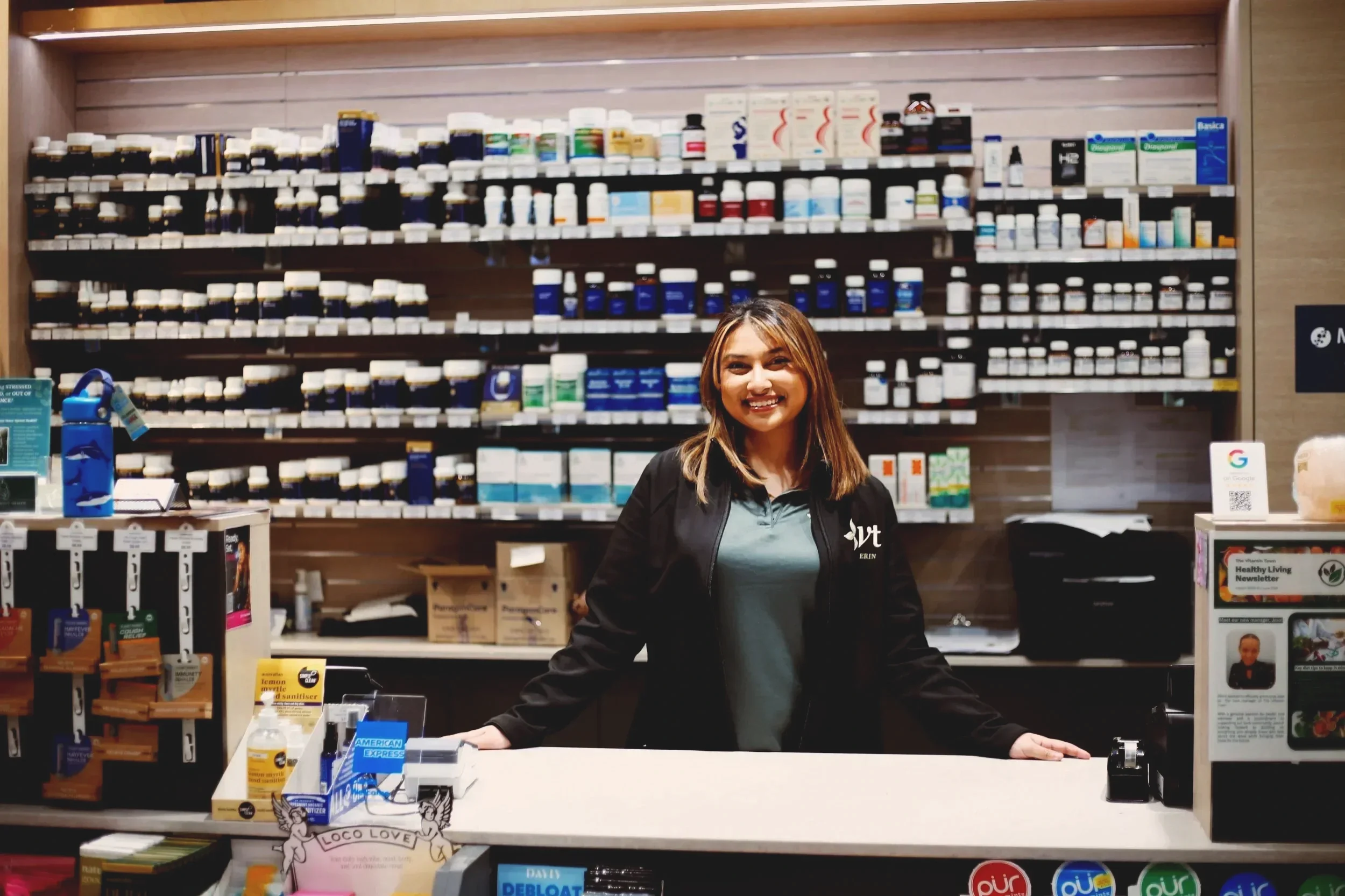 Smiling pharmacy employee standing behind a counter with shelves of vitamins and supplements.