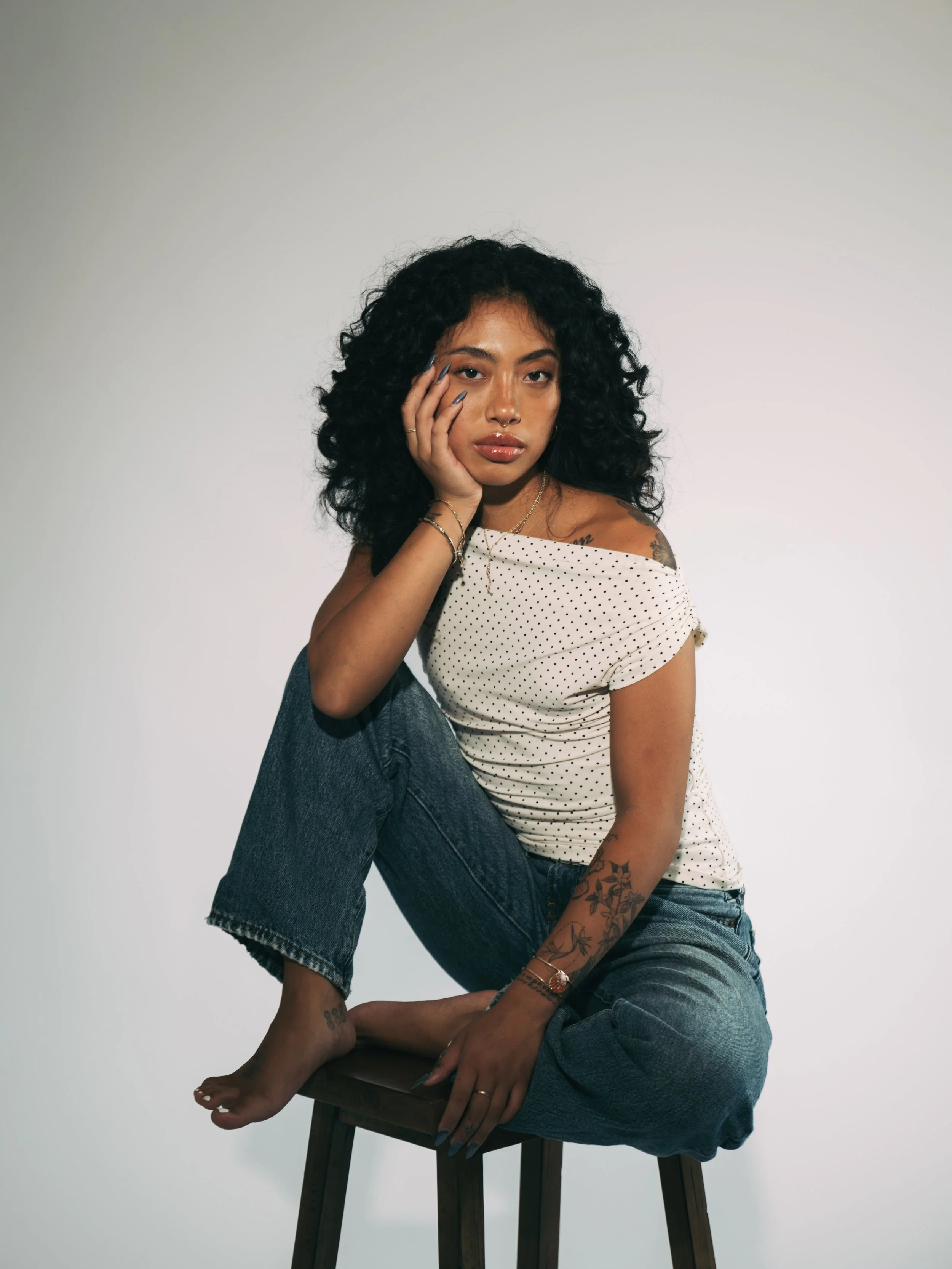 Tattooed woman with curly hair sitting barefoot on a wooden stool, wearing a polka-dot top and jeans against a plain background