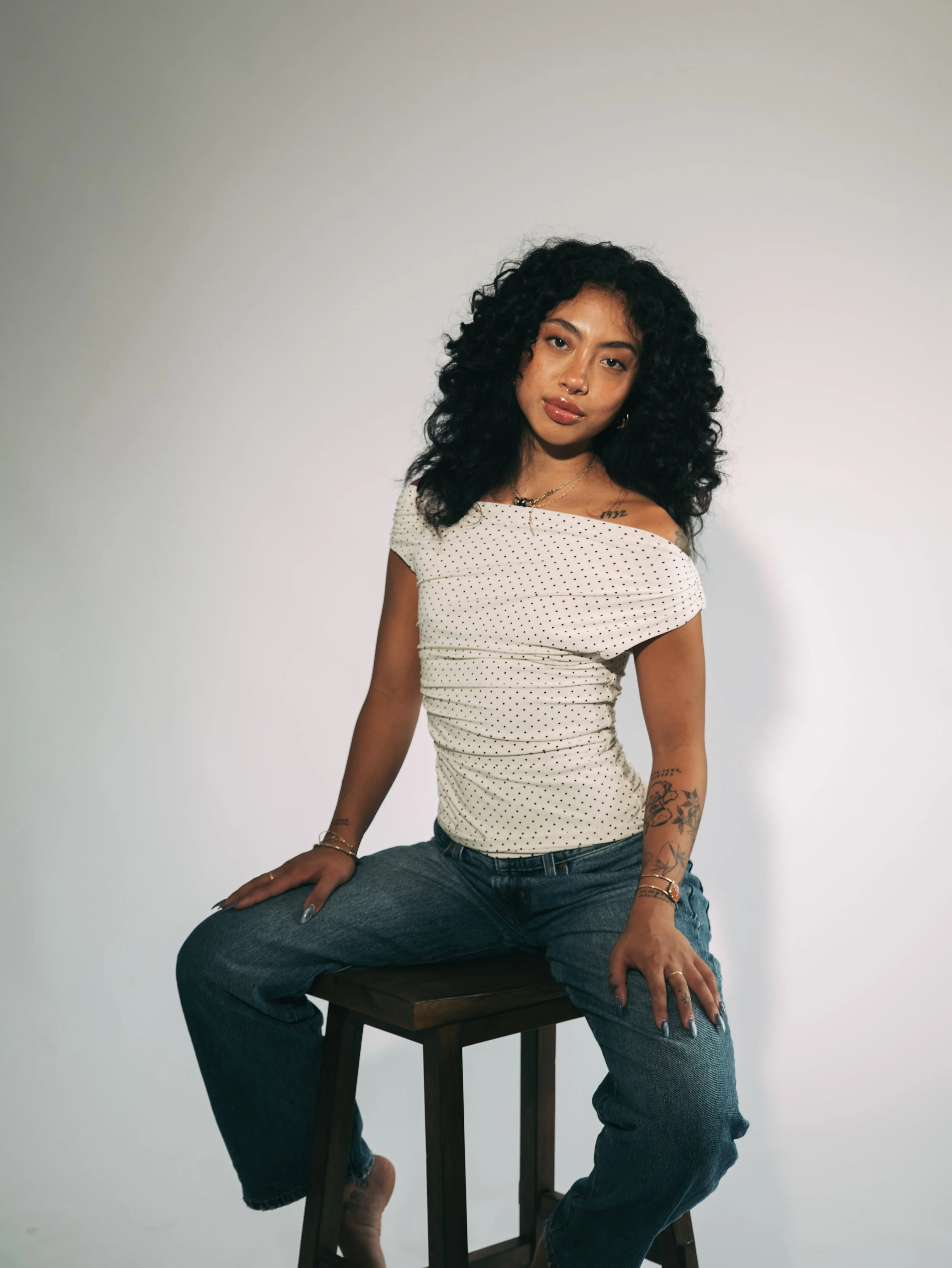 Tattooed woman with curly black hair sitting on a wooden stool, wearing a white polka-dot top and jeans.