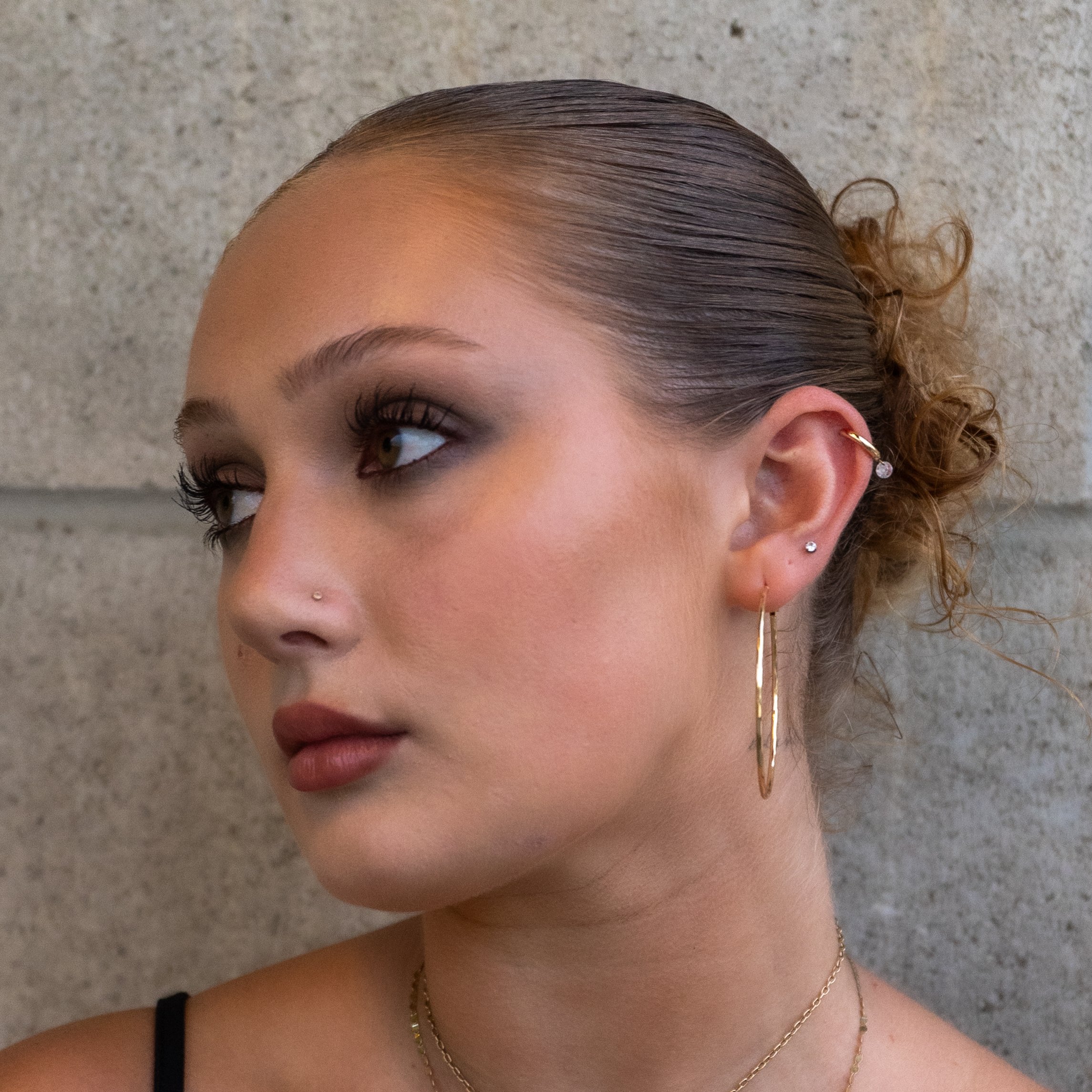 Close-up portrait of a woman with slicked-back hair, dramatic eye makeup, a nose stud, and layered gold earrings.