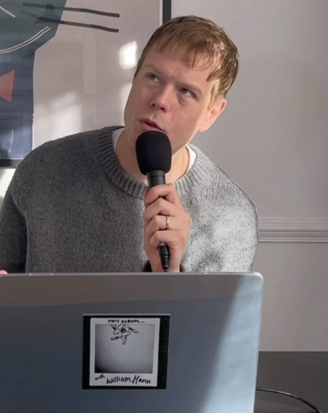 Man in a gray sweater holding a microphone behind a laptop labeled “My Album”