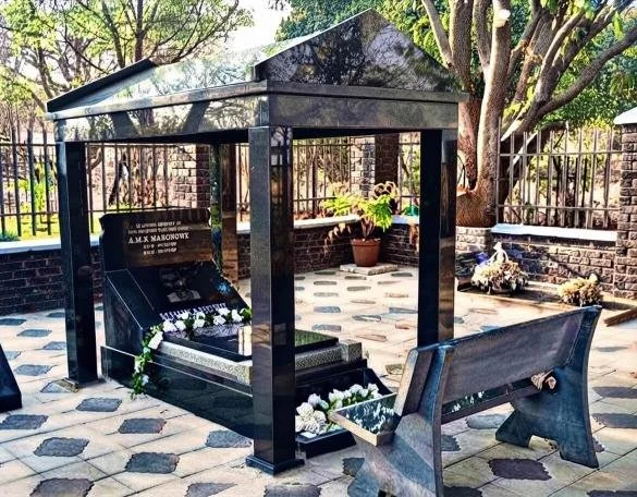 Outdoor memorial featuring a black granite tomb beneath a metal canopy, surrounded by flowers, trees, and stone paving.
