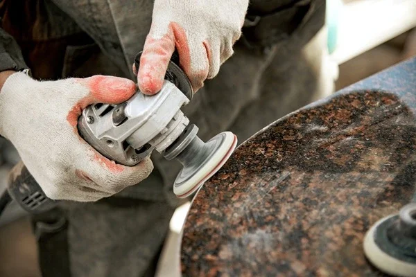 Gloved worker using an angle grinder to polish a granite stone surface