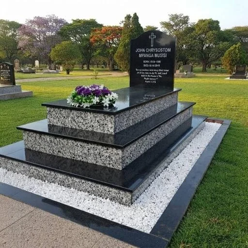 Polished black-and-white granite grave monument for John Chrysostom Mpanga-Musiisi in a landscaped cemetery, decorated with purple and white flowers.