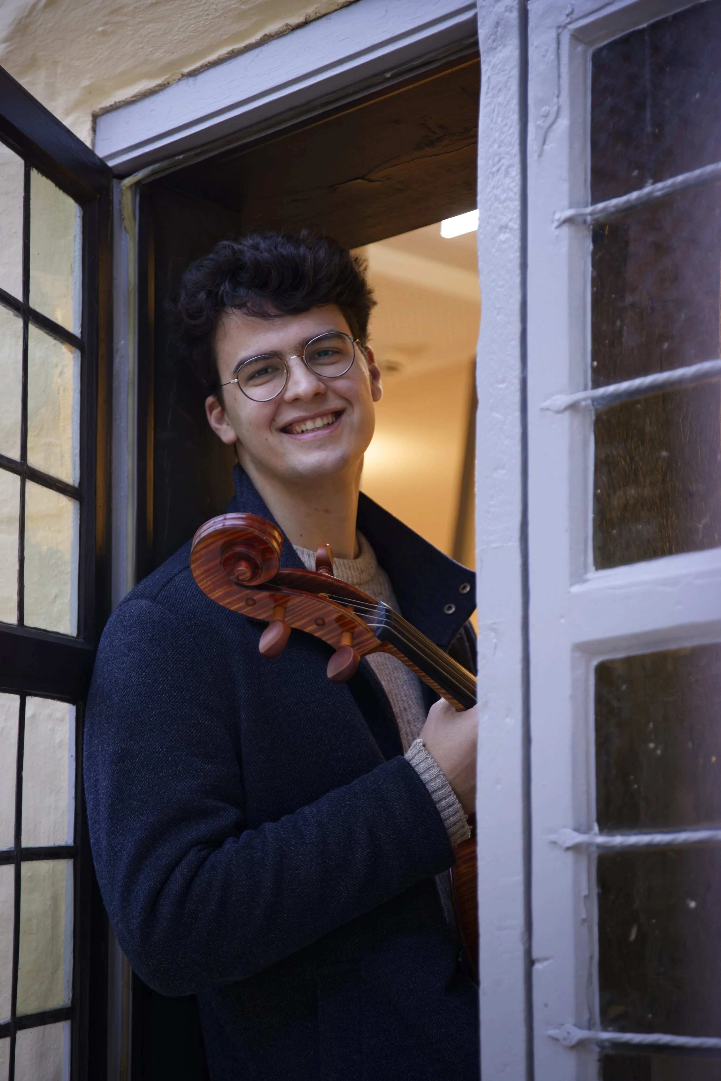 Lächelnder Musiker mit Brille und Cello steht in einem geöffneten Fenster.