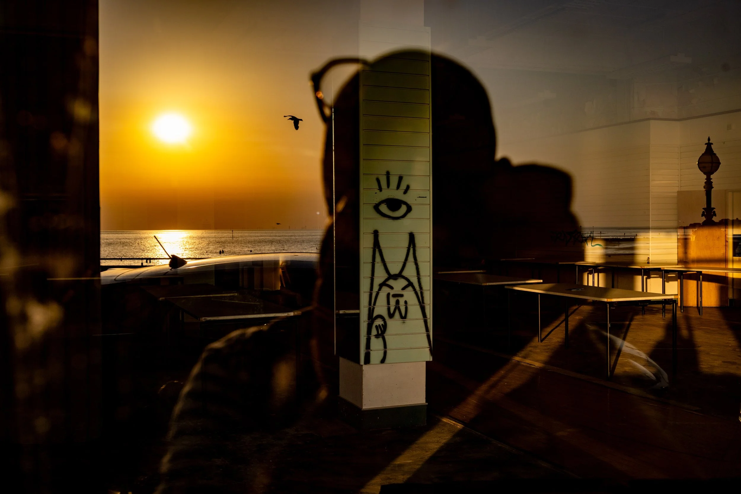 Sunset over the ocean viewed through a reflective seaside building, with a silhouetted bird, graffiti-covered column, tables, and lamp post. Photo by Jon Rees.