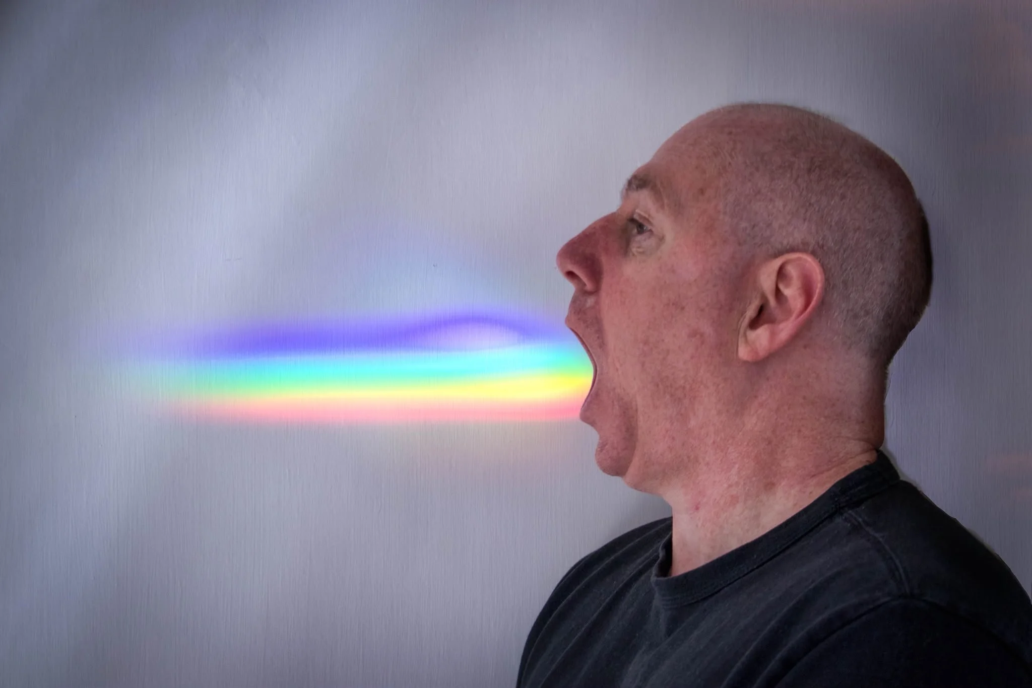 Side-profile of artist Jon Rees exhaling a colourful rainbow beam against a grey background