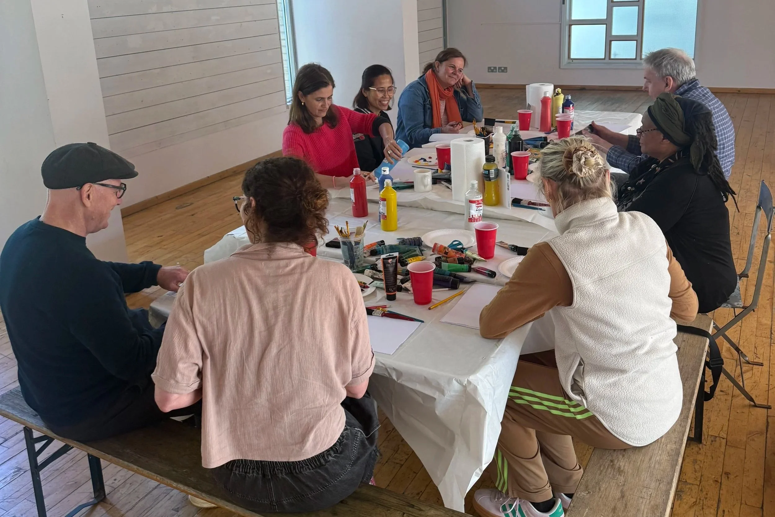 Group of adults participating in a painting workshop around a table covered with art supplies, paper, brushes, and colorful paints.