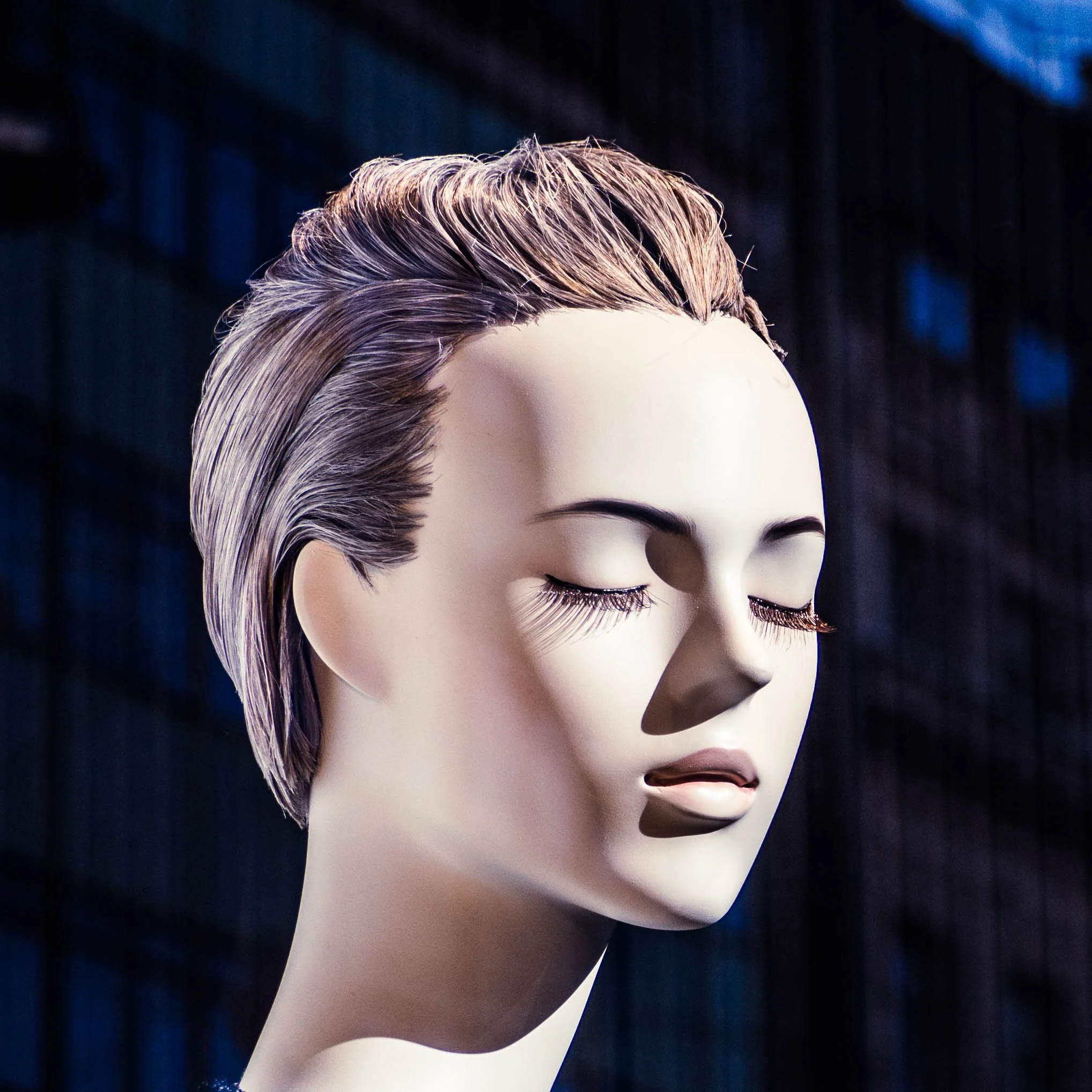 Close-up of a mannequin head with short, swept-back blonde hair and closed eyes against a dark background. Photo by Jon Rees.