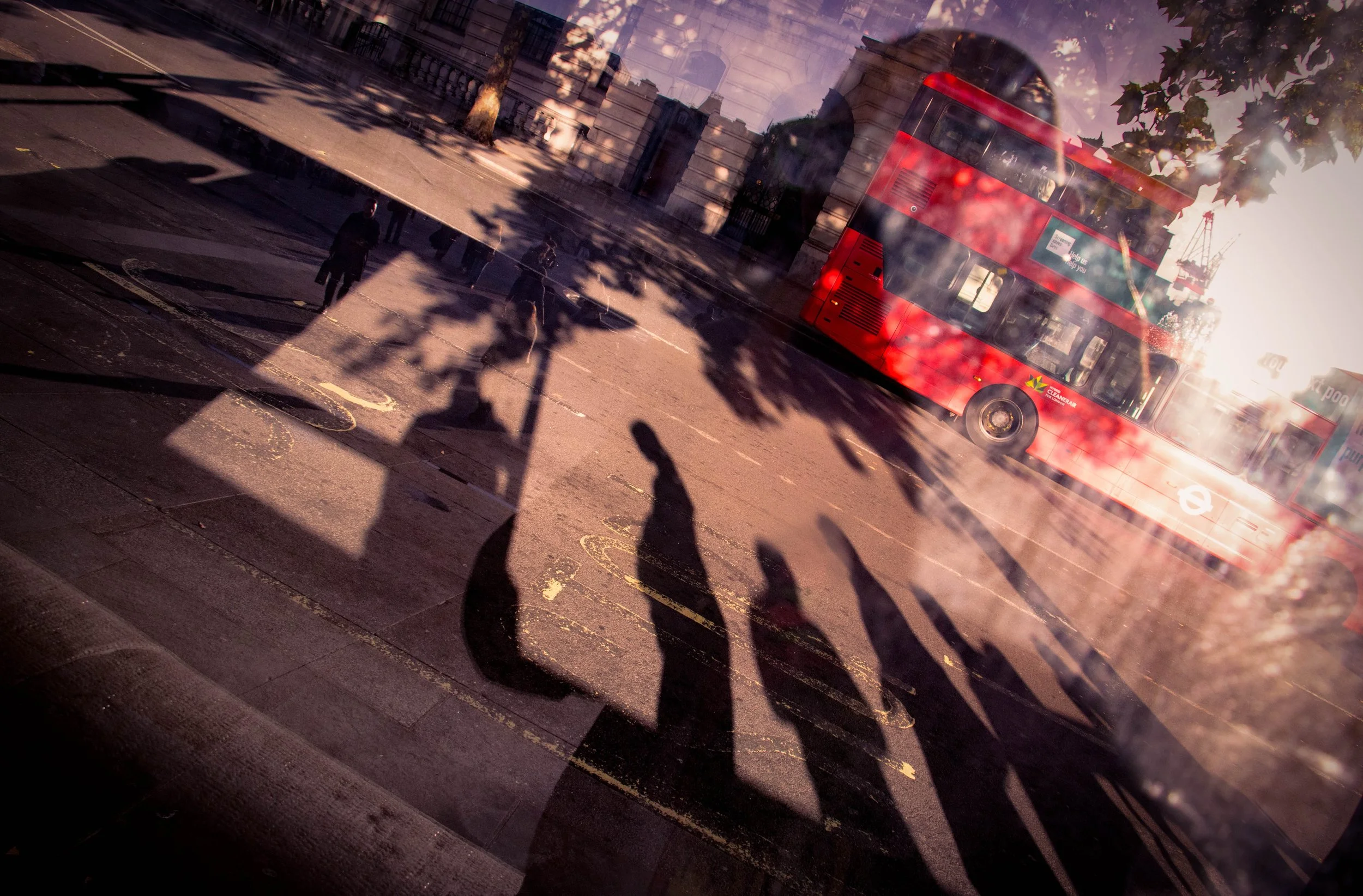 Red double-decker bus passing pedestrians and long shadows on a sunlit city street. Photo by Jon Rees.