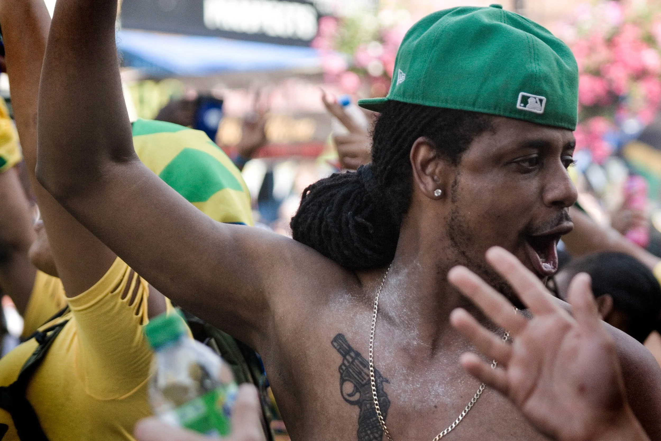 Shirtless man with dreadlocks and a green baseball cap cheering in a crowded outdoor celebration. Photo by Jon Rees.