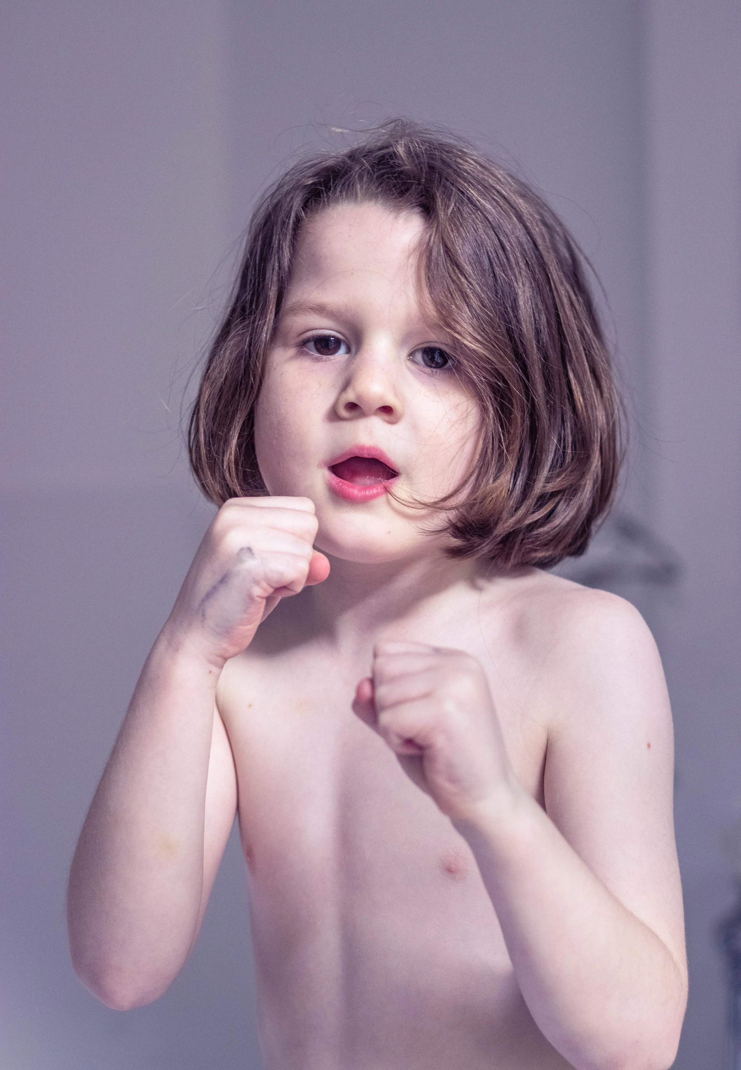 Close-up of a young child with brown hair and an open mouth, raising both fists against a gray background. Photo by Jon Rees.