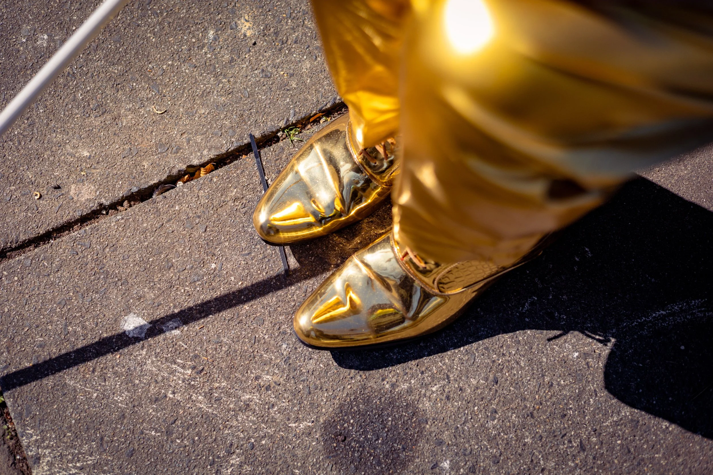 Close-up of reflective gold platform shoes on a sunlit asphalt street. Photo by Jon Rees.