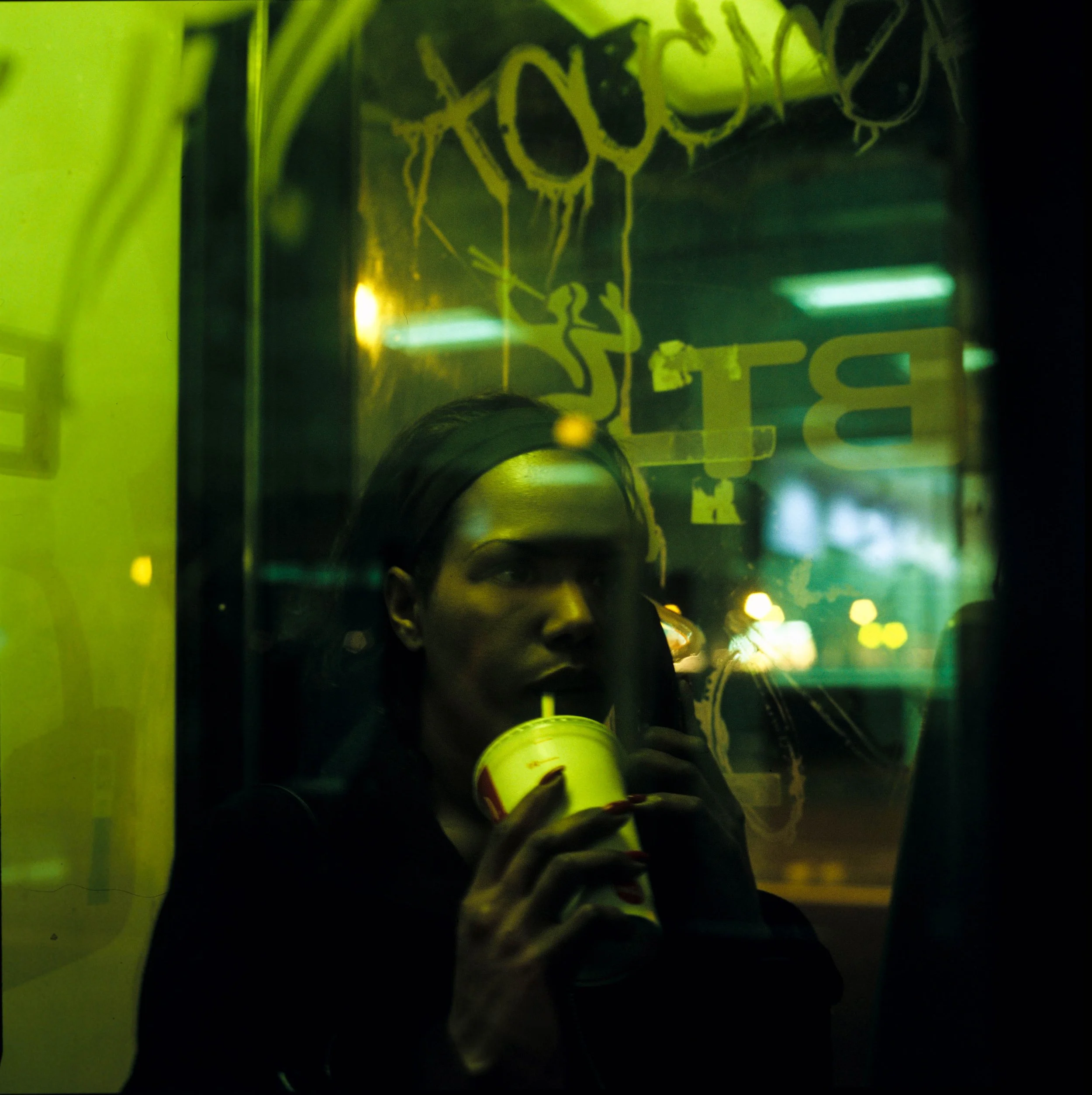 Person wearing a black hoodie and holding a takeaway coffee cup in a dimly lit, green-tinted setting.
