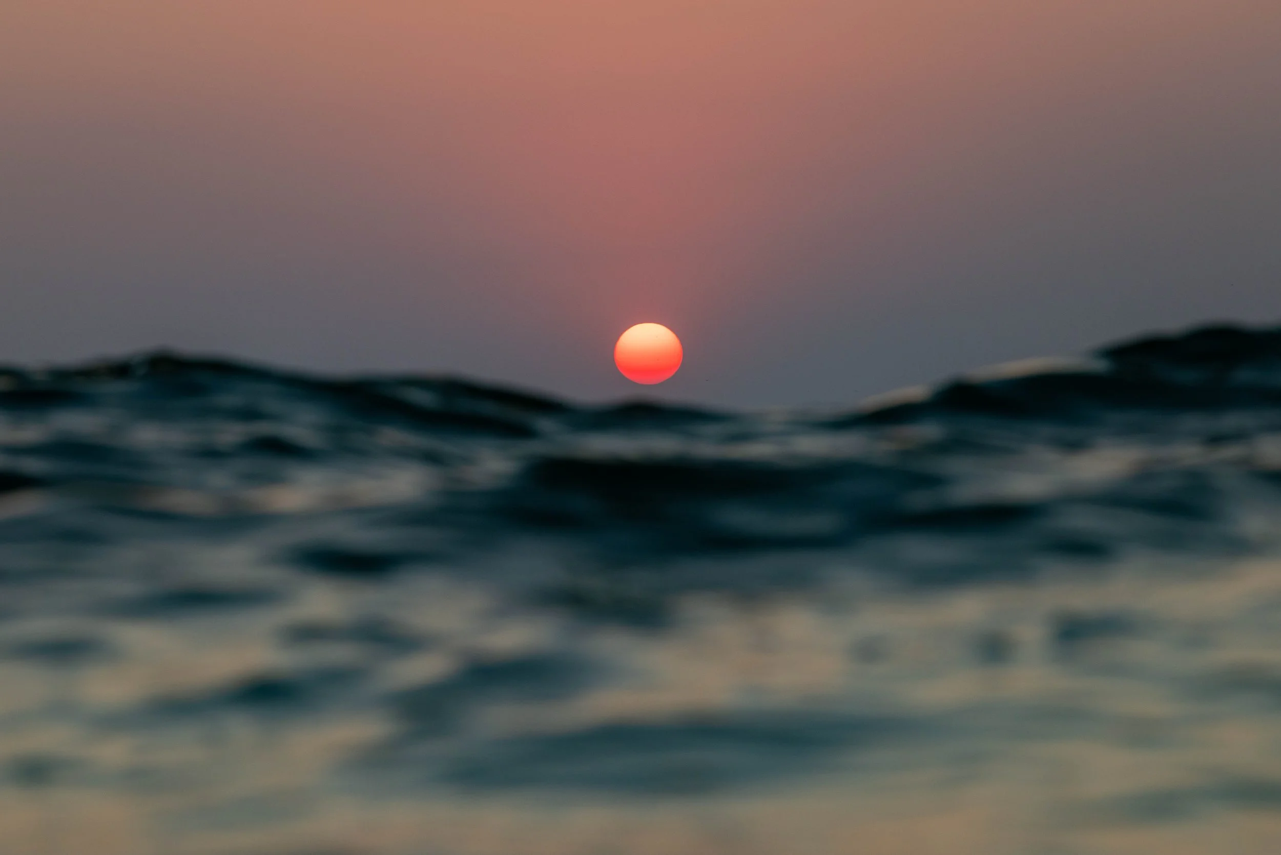 Red-orange sun setting over blurred ocean waves beneath a dusky sky. Photo by Jon Rees.