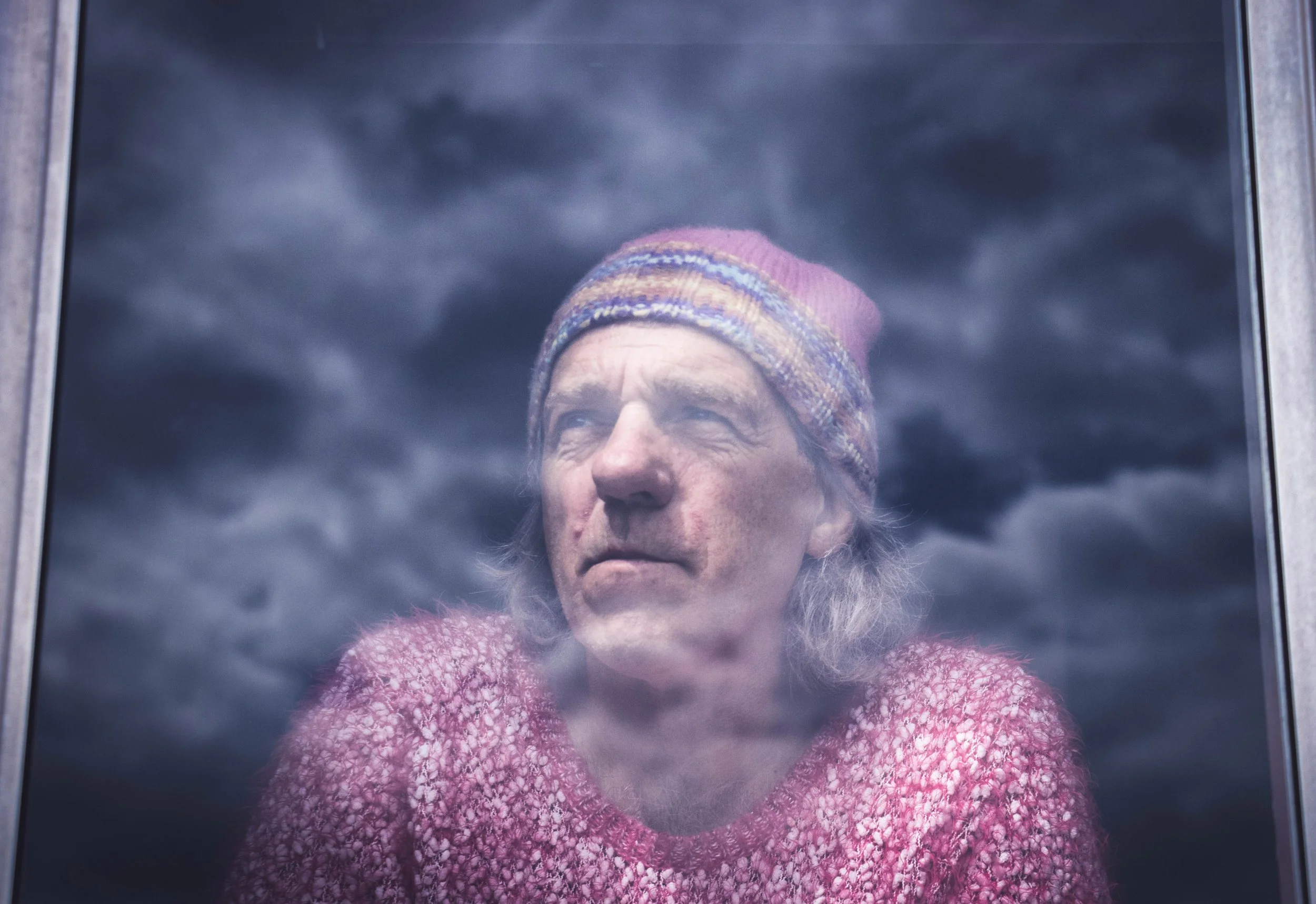 Close-up portrait of an older woman wearing a multicolored knit beanie and pink sweater, viewed through a window. Photo by Jon Rees.