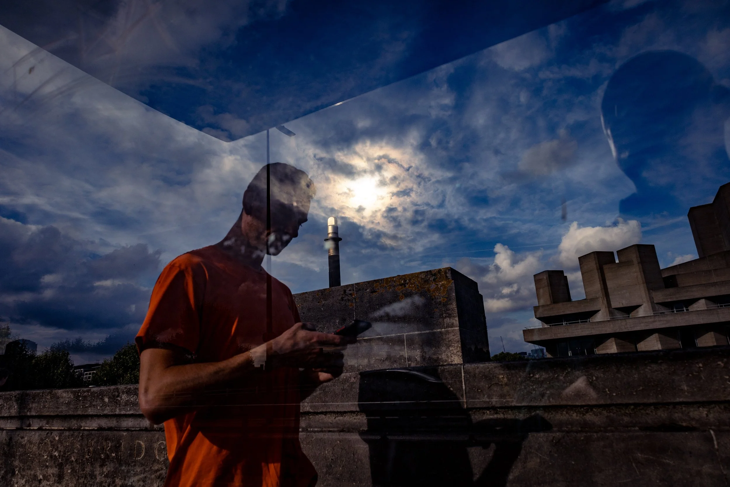 Man in an orange shirt using a smartphone near a concrete industrial building under a dramatic cloudy sky. Photo by Jon Rees.