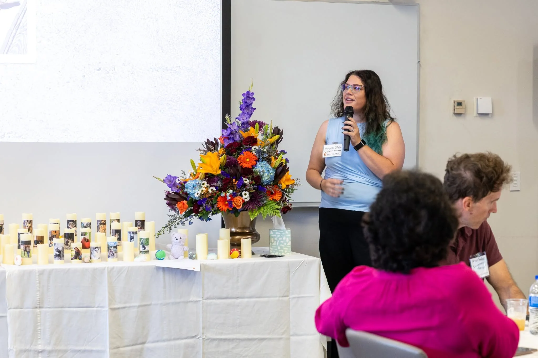 Woman speaking into a microphone beside a colorful floral arrangement and memorial candles at an indoor gathering.