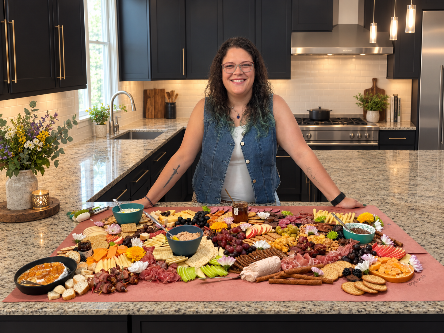 Smiling woman standing behind an elaborate charcuterie board with meats, cheeses, fruit, crackers, dips, and flowers in a modern black kitchen
