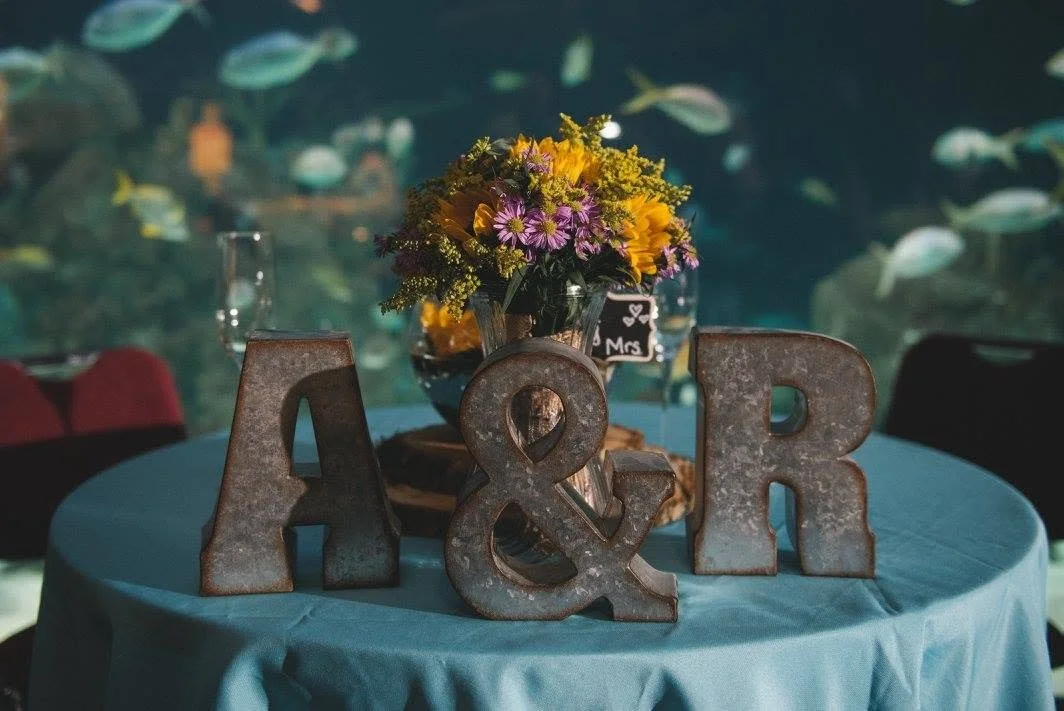 Rustic metal A&R initials with an ampersand on a blue table, floral centerpiece, and aquarium fish in the background