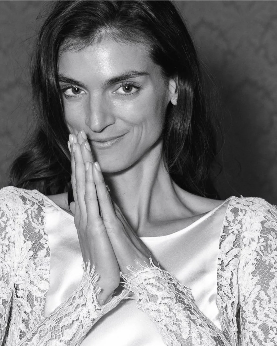 Black-and-white portrait of a smiling woman with long dark hair, hands pressed together, wearing a lace-trimmed top
