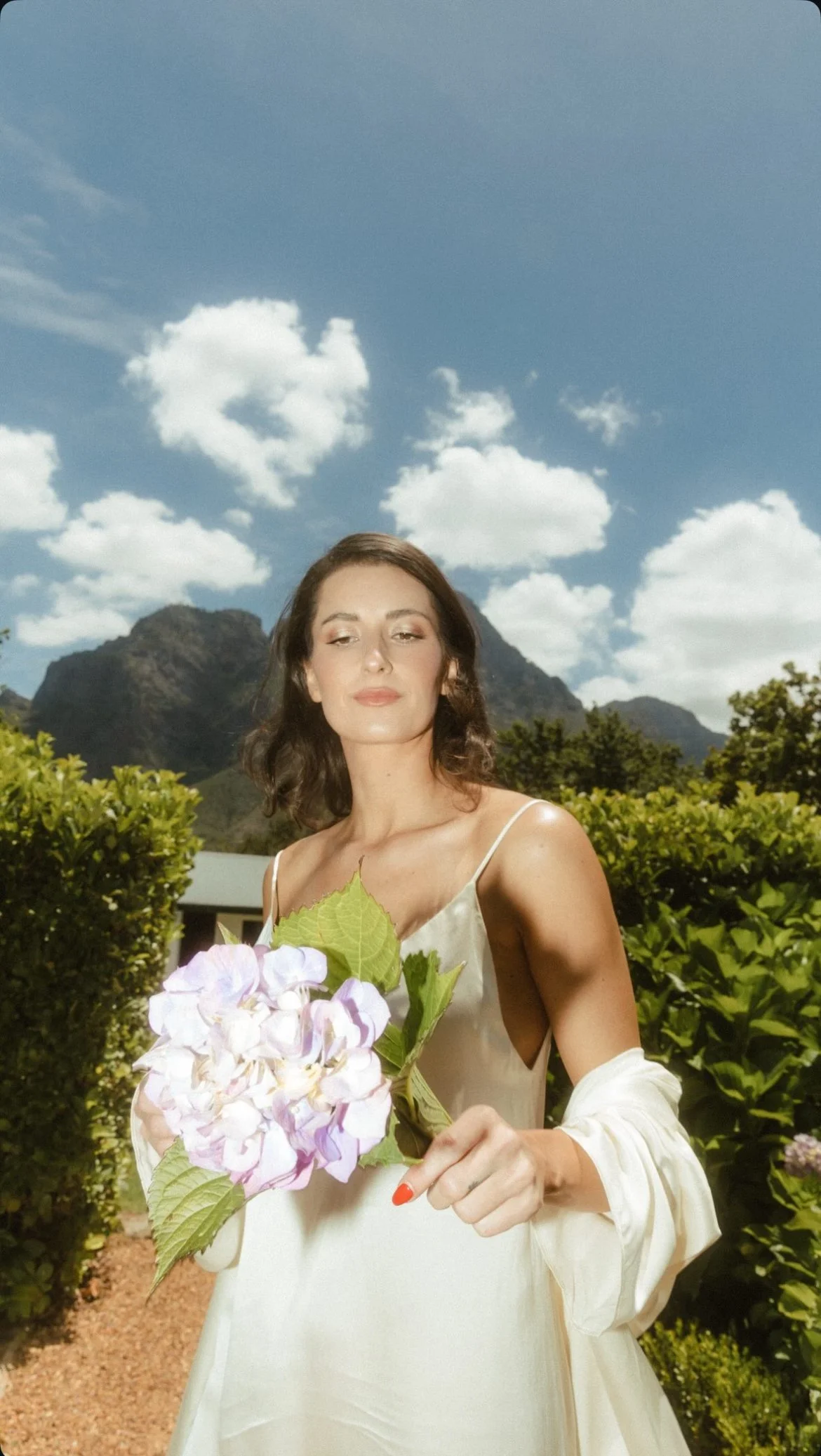 Woman in a white dress holding a bouquet of hydrangeas in a tropical garden with mountains and clouds behind her