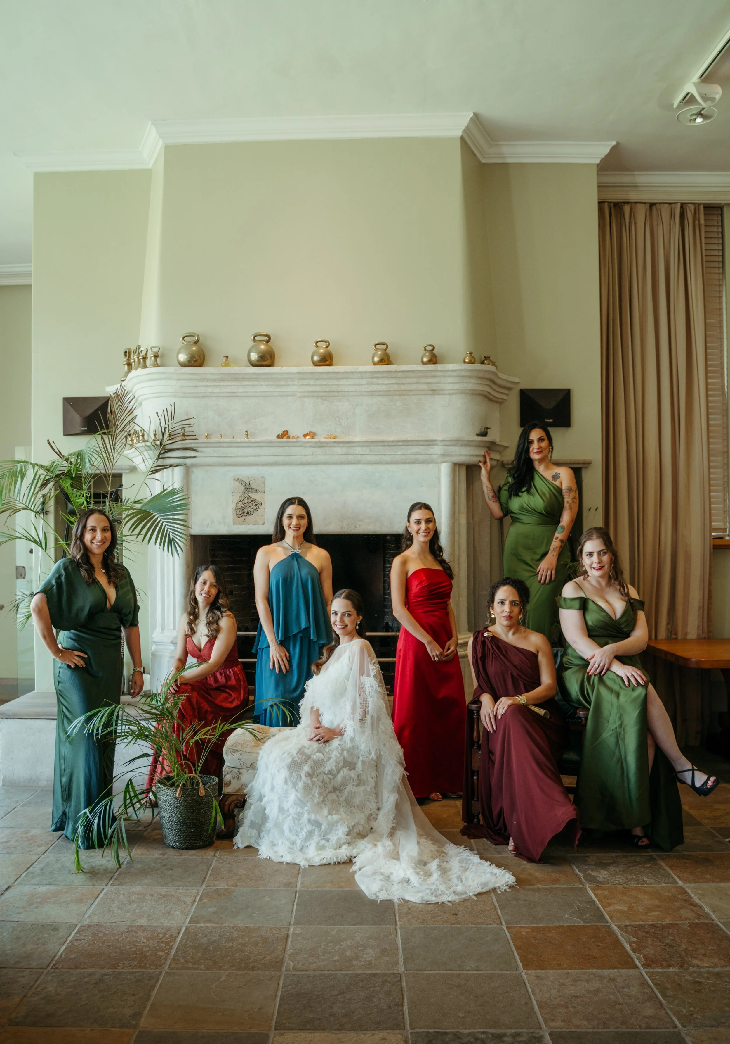 Group portrait of women in colorful formal dresses gathered around an ornate fireplace, with a bride in a white gown seated in front.