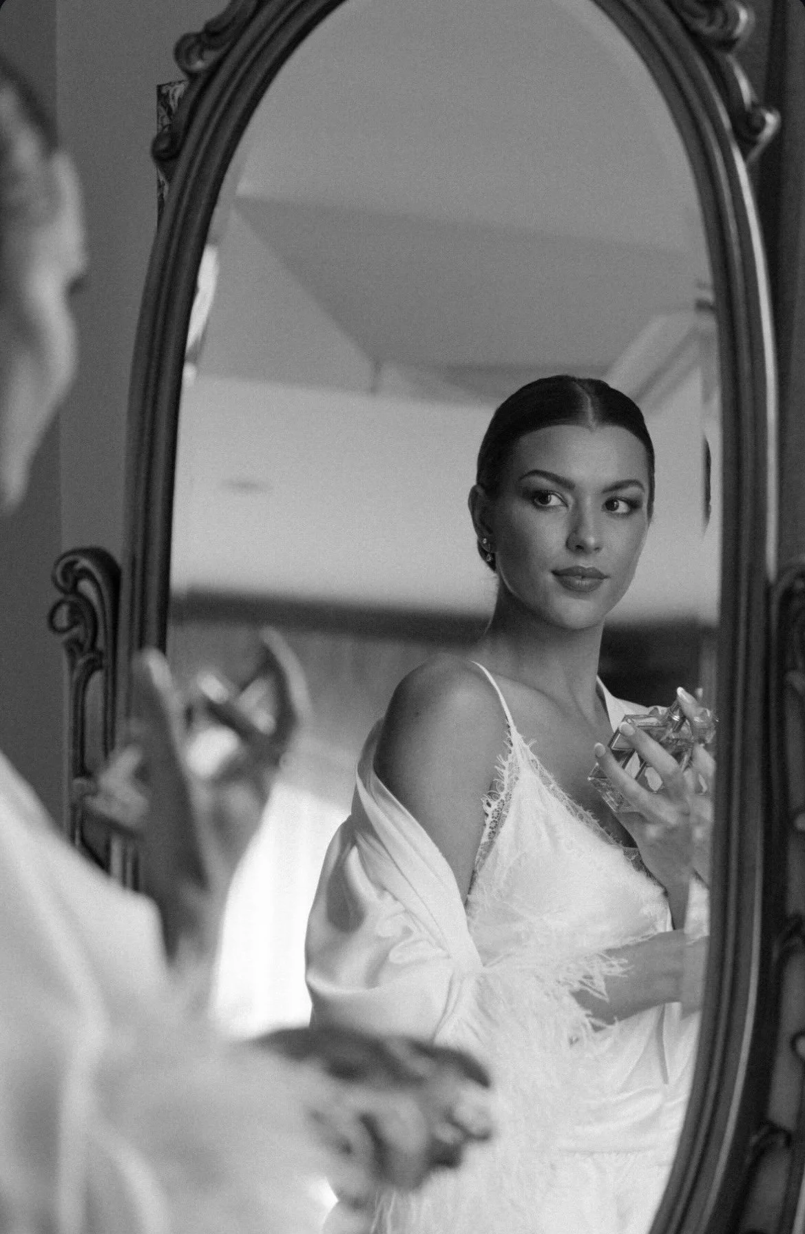 Black-and-white portrait of a woman in a lace-trimmed dress holding a perfume bottle while looking into an ornate mirror