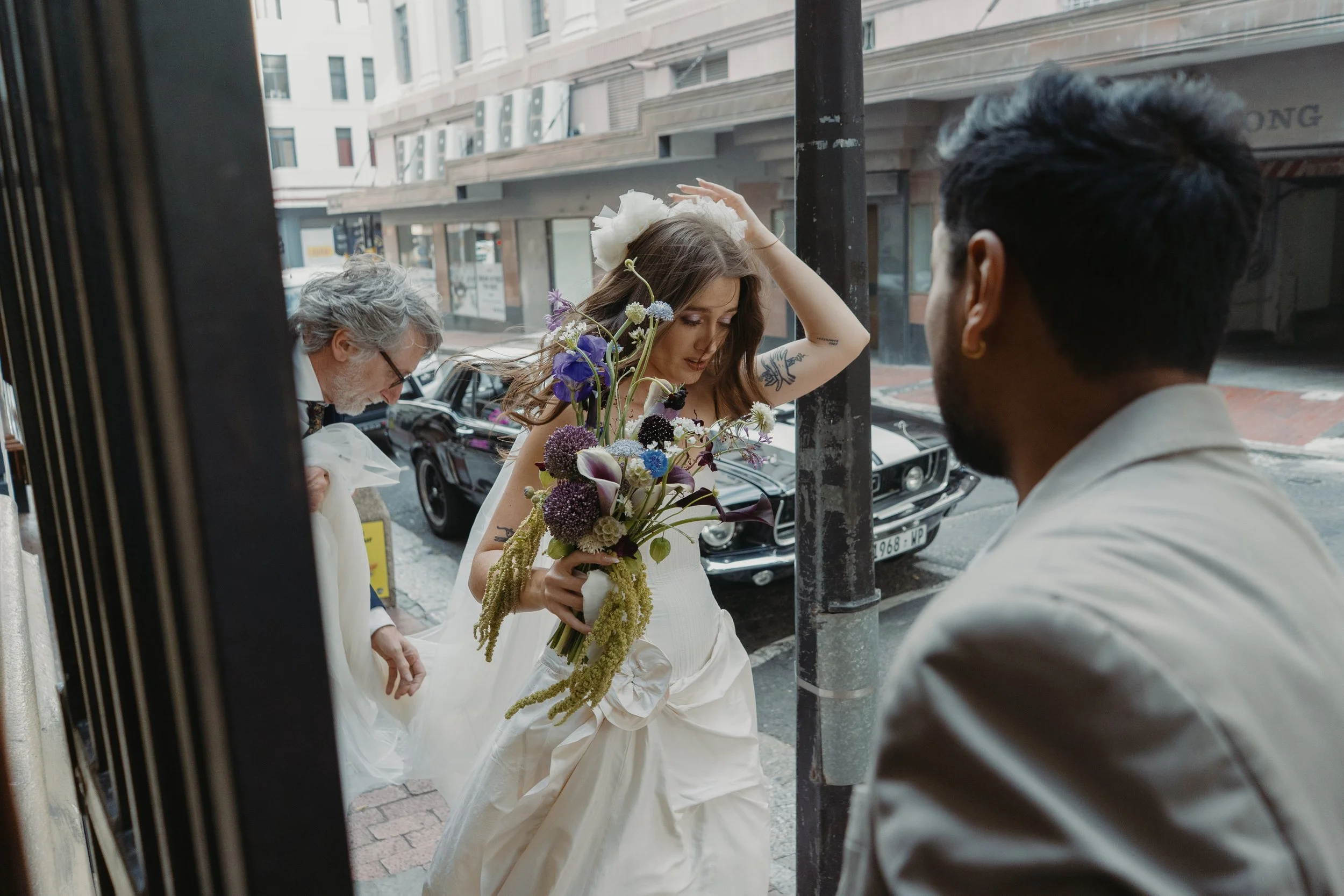 Bride in a white dress and floral headpiece holding a colorful bouquet outdoors with two people nearby