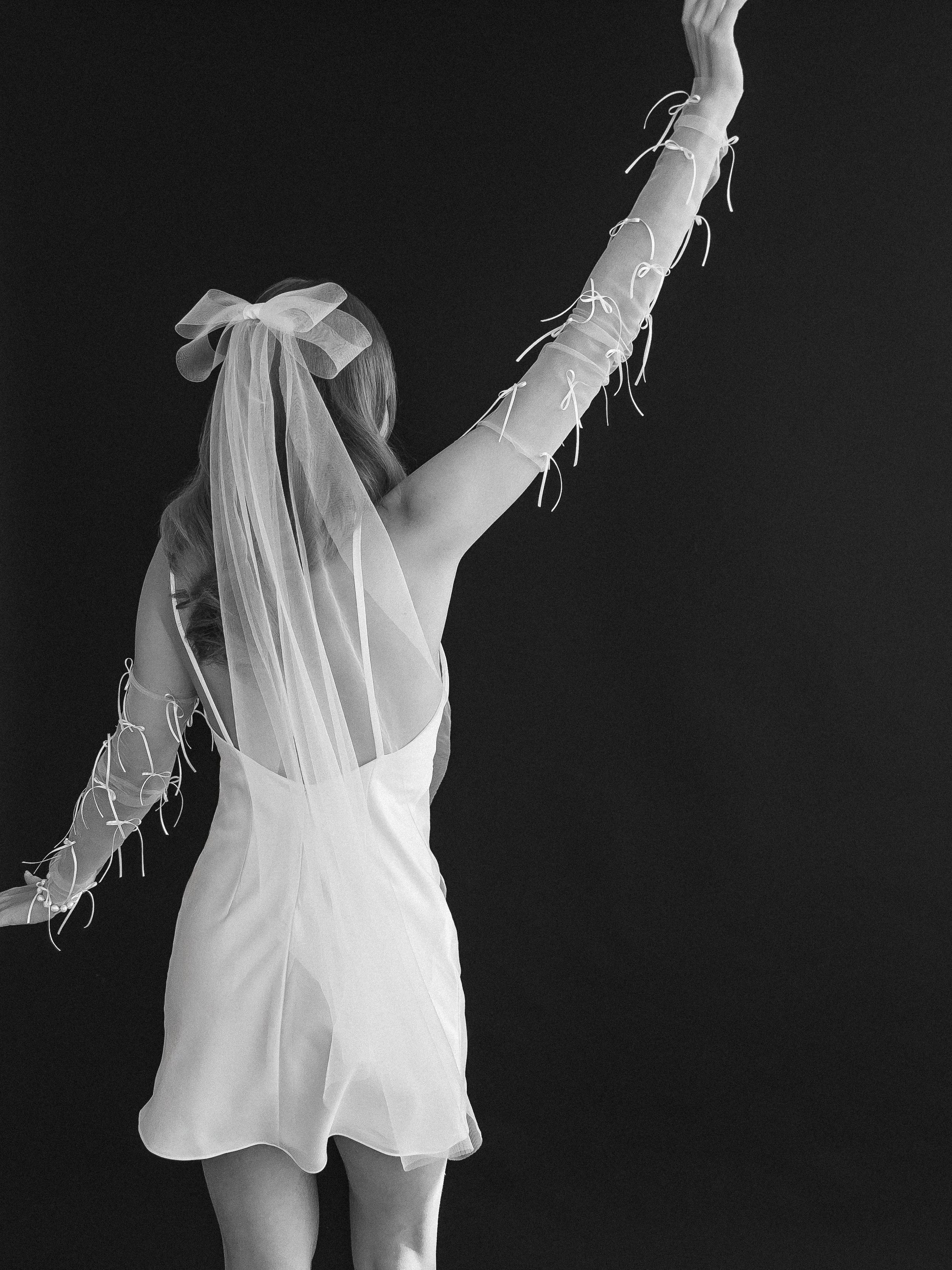 Black-and-white photo of a person in a flowing dress and large bow raising an arm decorated with ribbons against a dark background