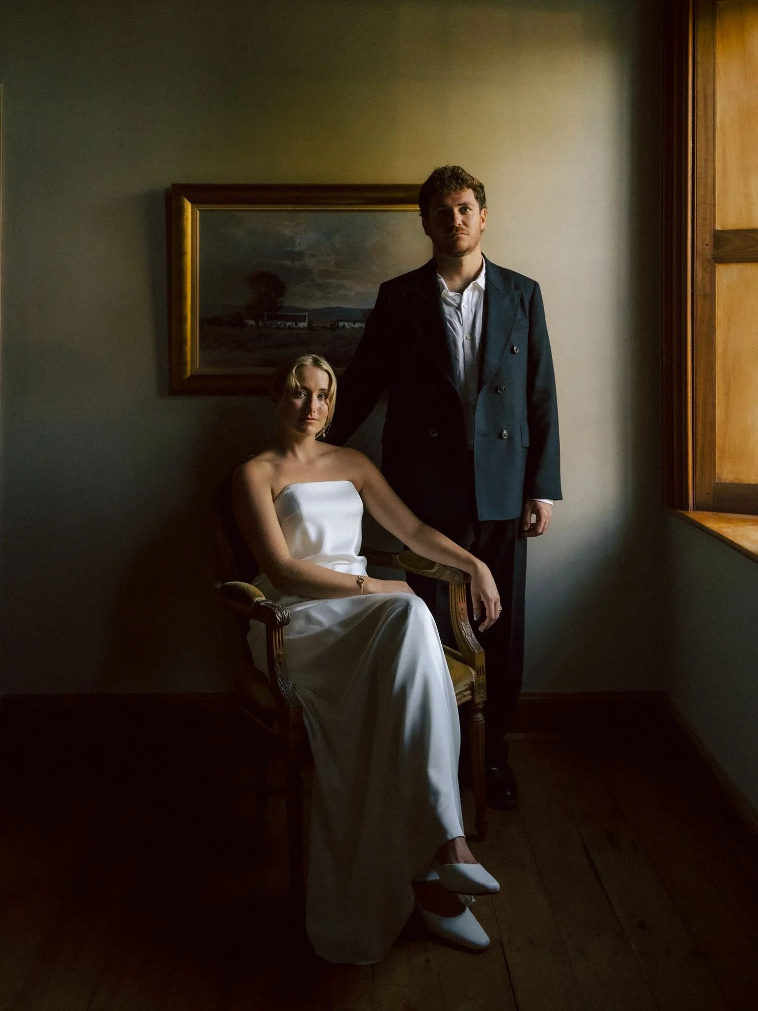 Woman in a white wedding dress seated in an ornate chair beside a man in a dark double-breasted suit, with a framed painting behind them.