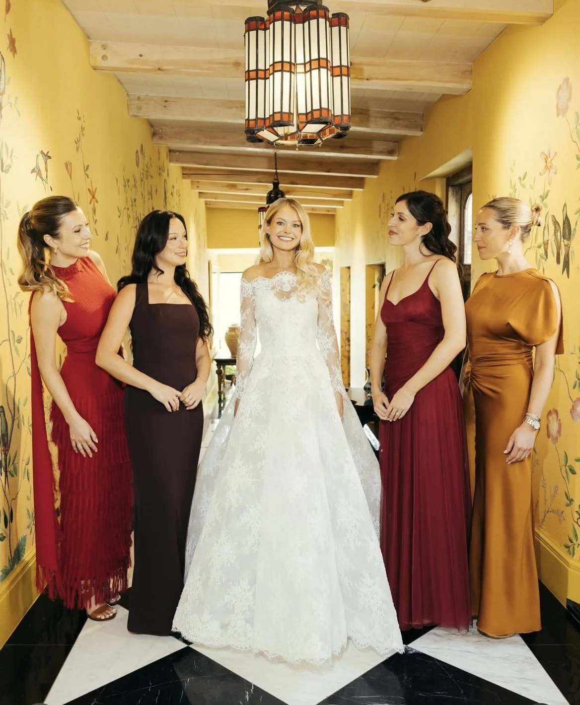 Smiling bride in a white lace wedding gown standing with four bridesmaids in colorful dresses in a floral hallway