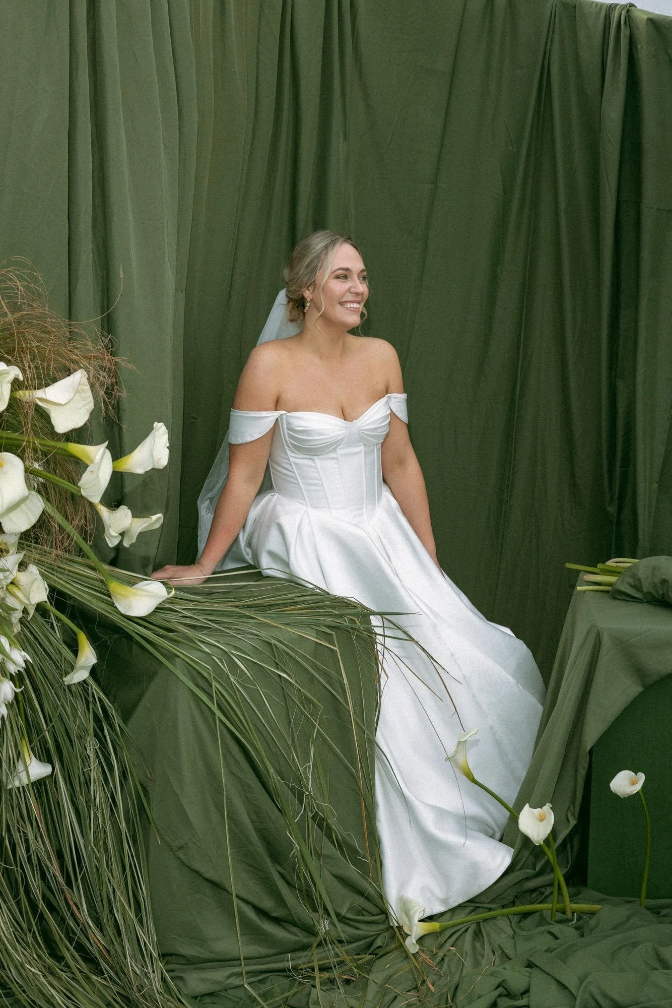 Smiling bride in an off-the-shoulder white wedding gown and veil surrounded by white calla lilies against a green draped backdrop