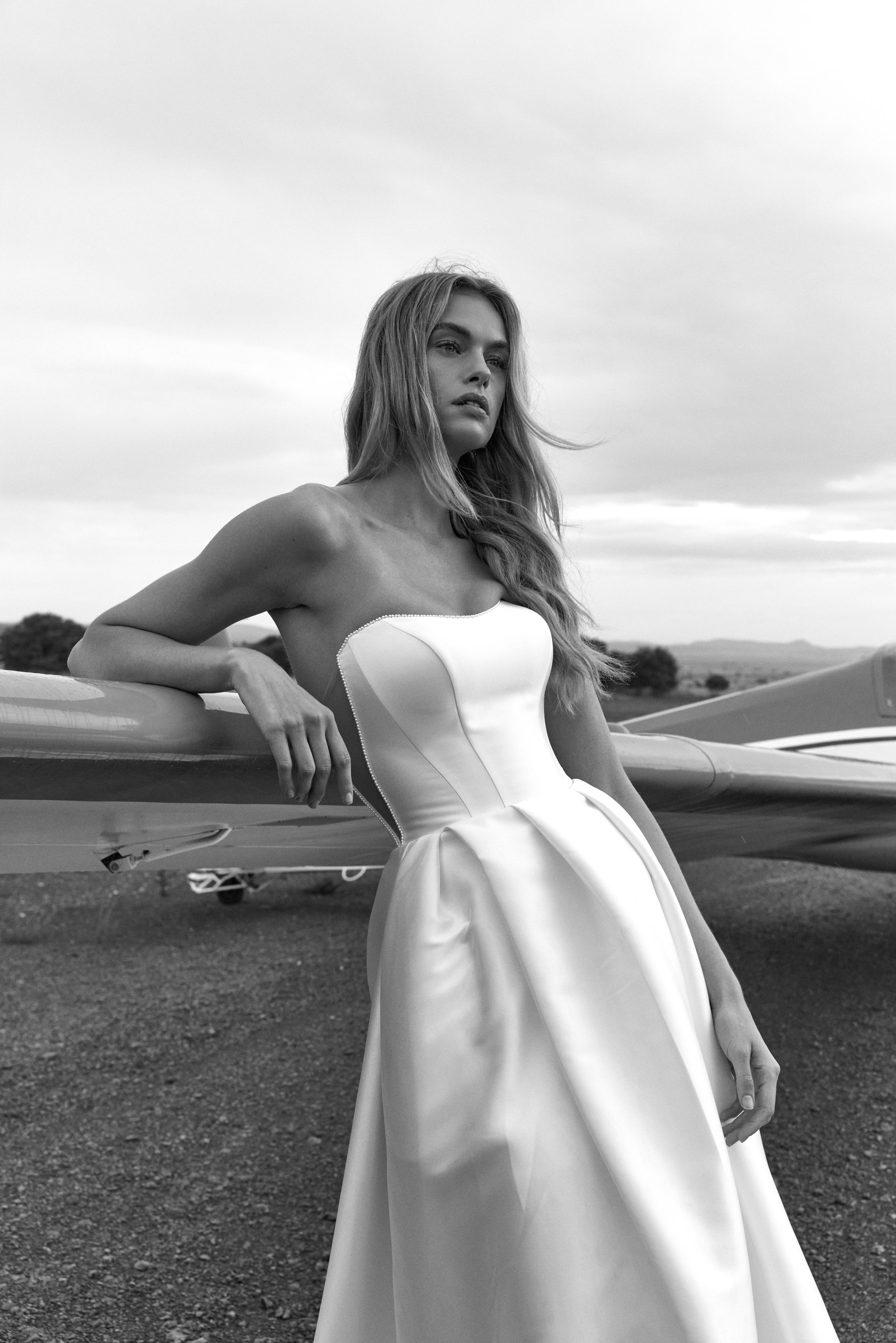 Black-and-white portrait of a woman in a white dress posing beside a vintage car outdoors