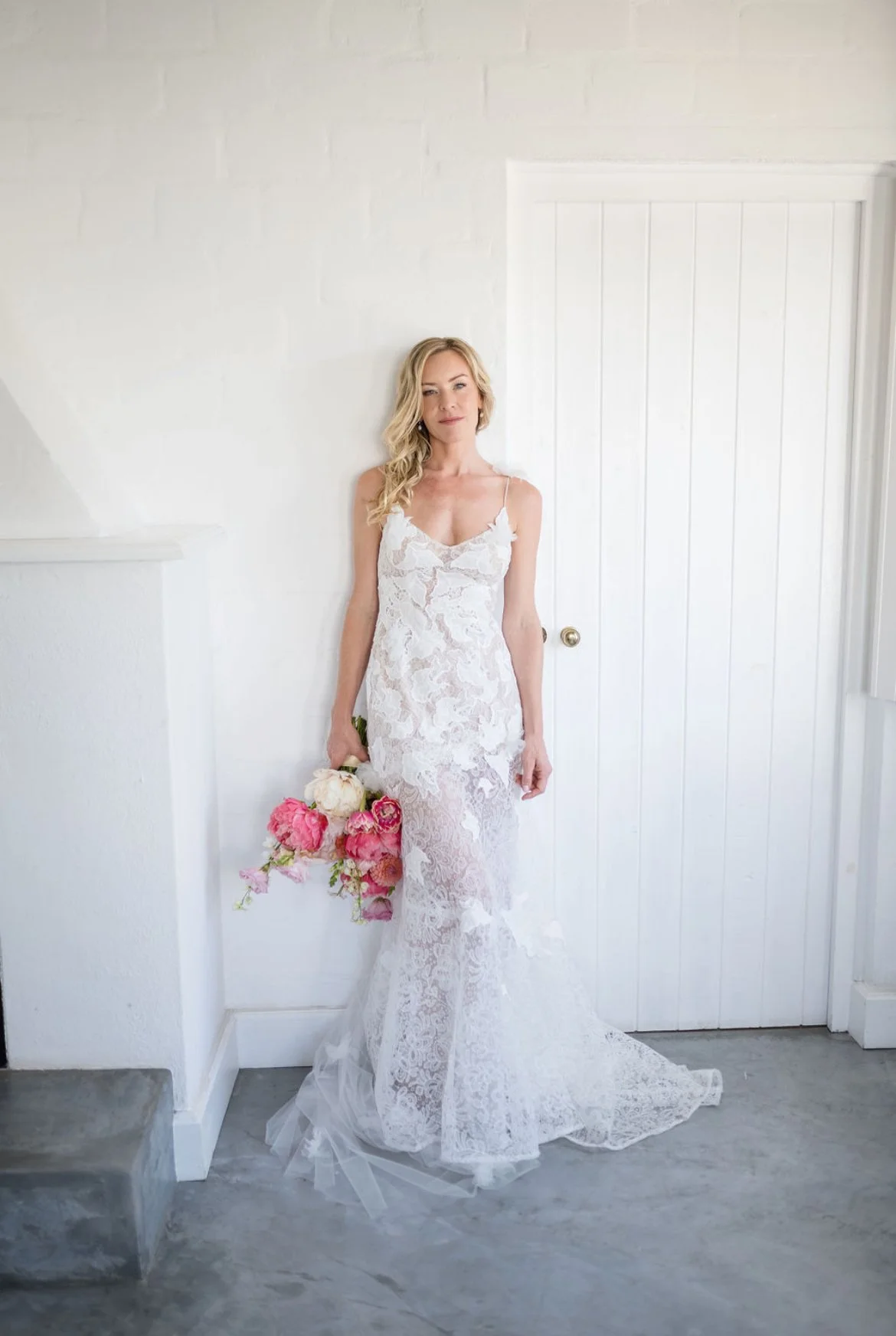 Woman in a white lace wedding dress holding a bouquet of pink and white flowers indoors