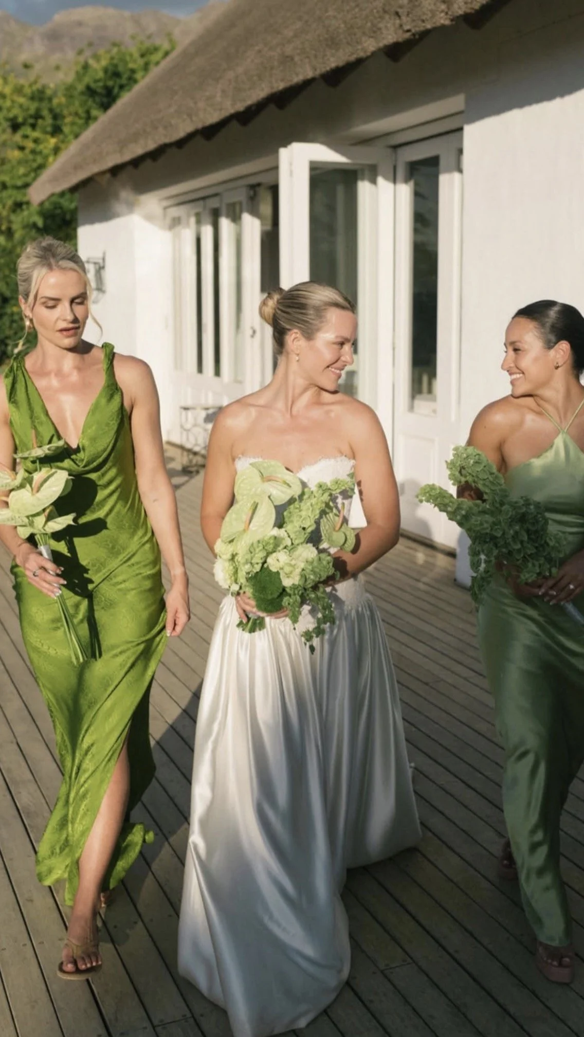 Bride and two bridesmaids in green dresses holding bouquets outside a thatched-roof venue