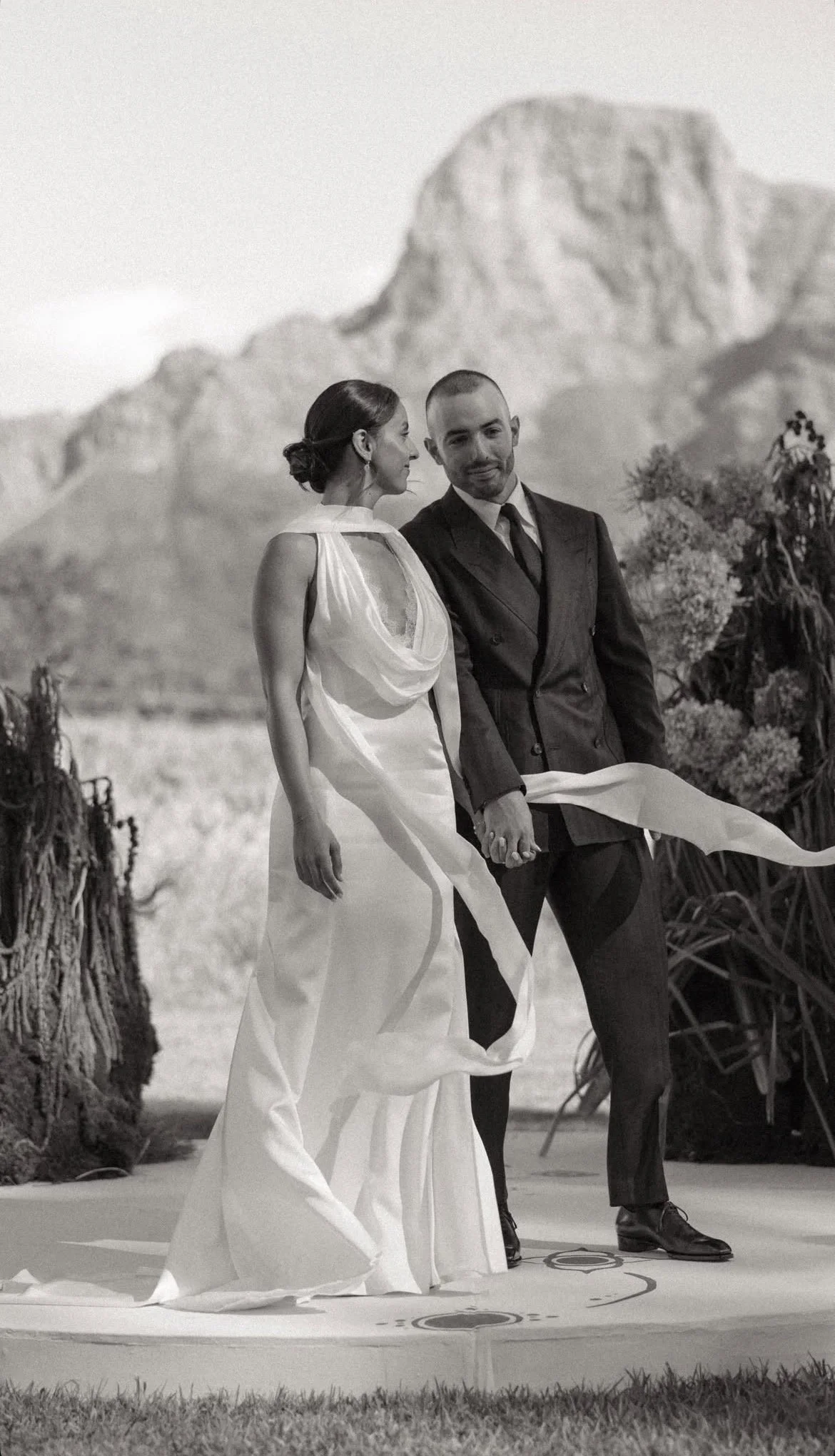 Black-and-white wedding portrait of a bride and groom holding hands outdoors with a mountain backdrop