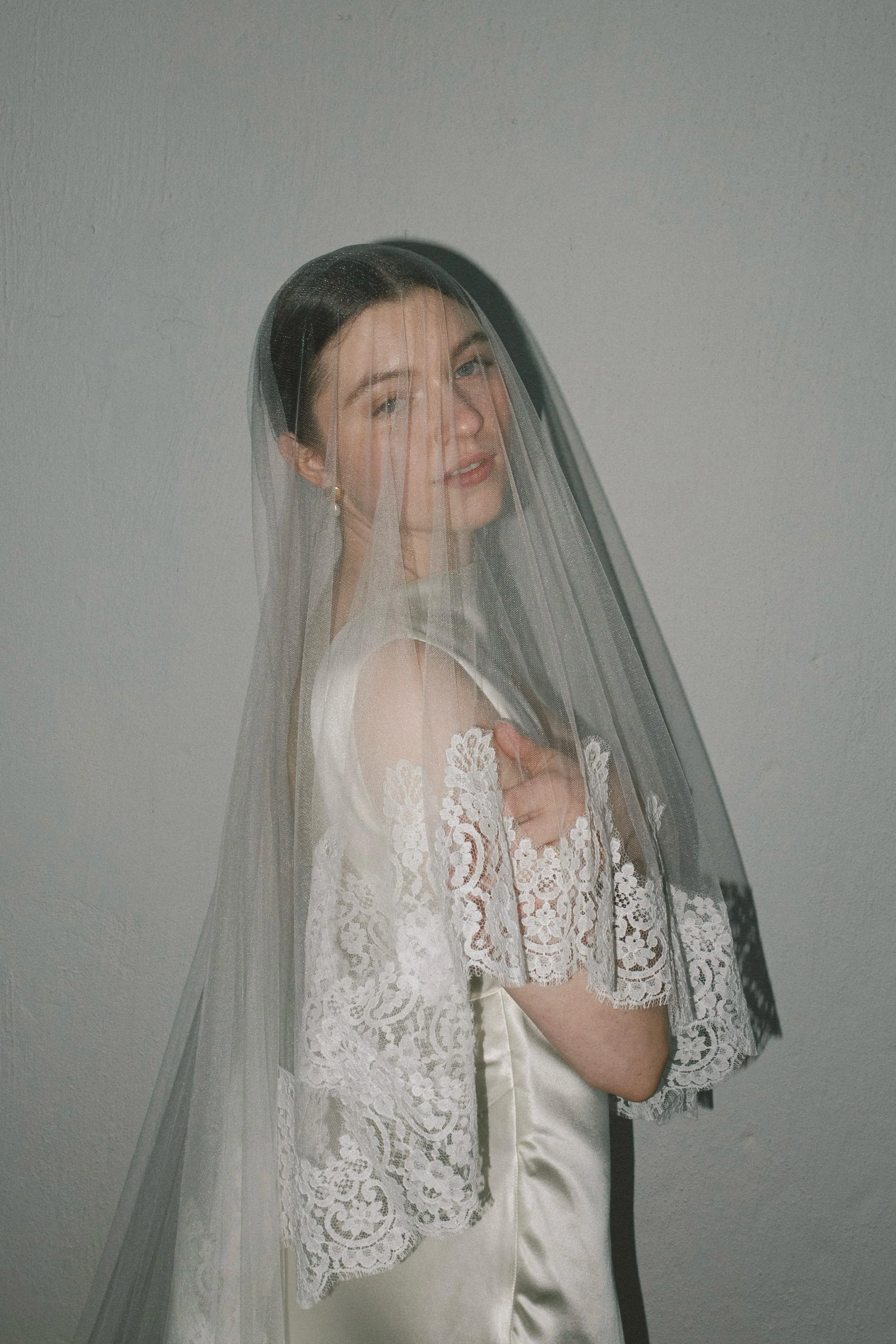 Smiling bride in a lace wedding dress and sheer veil posing against a gray wall