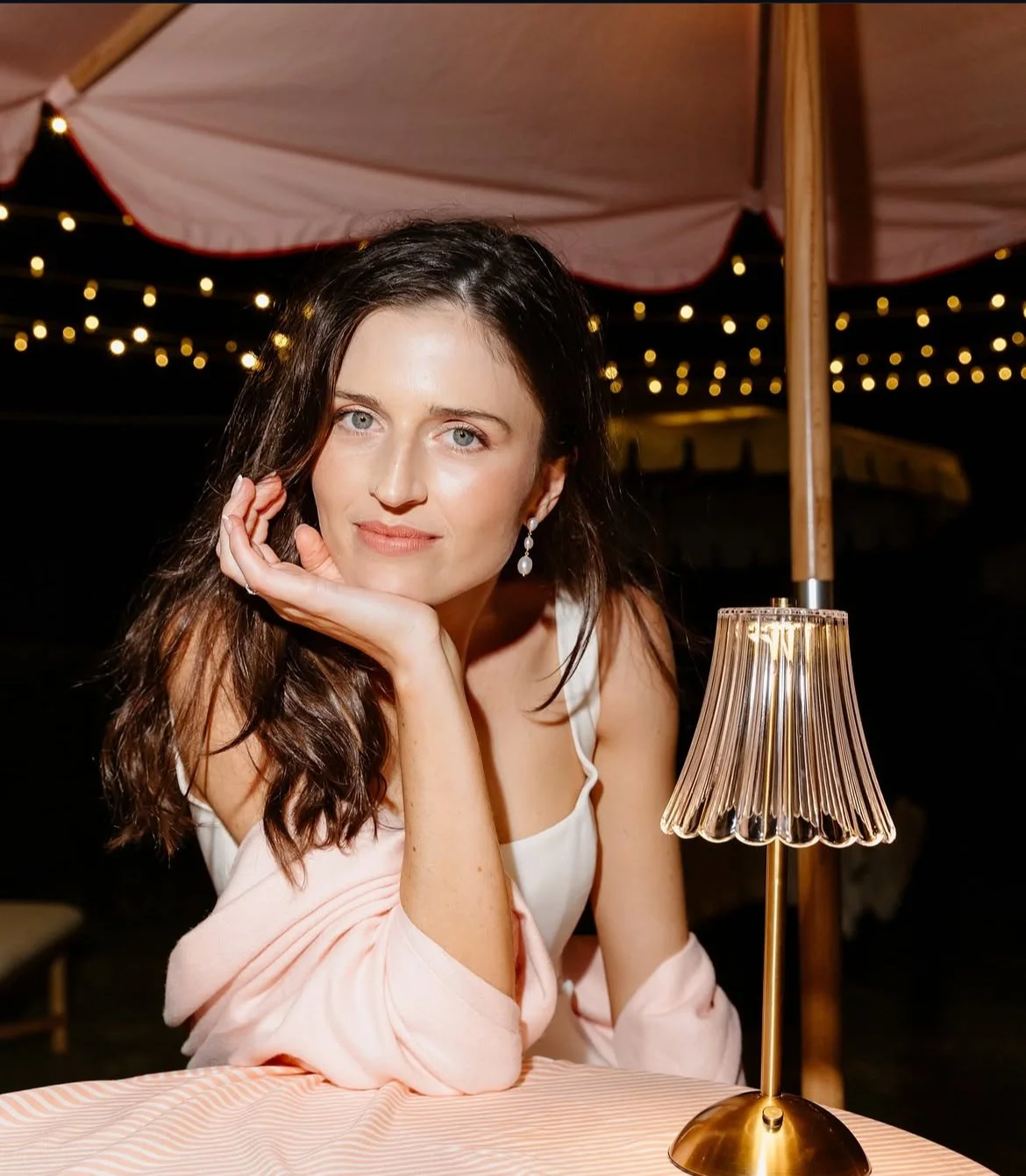 Woman in a white dress and pink shawl posing beside a vintage table lamp under string lights