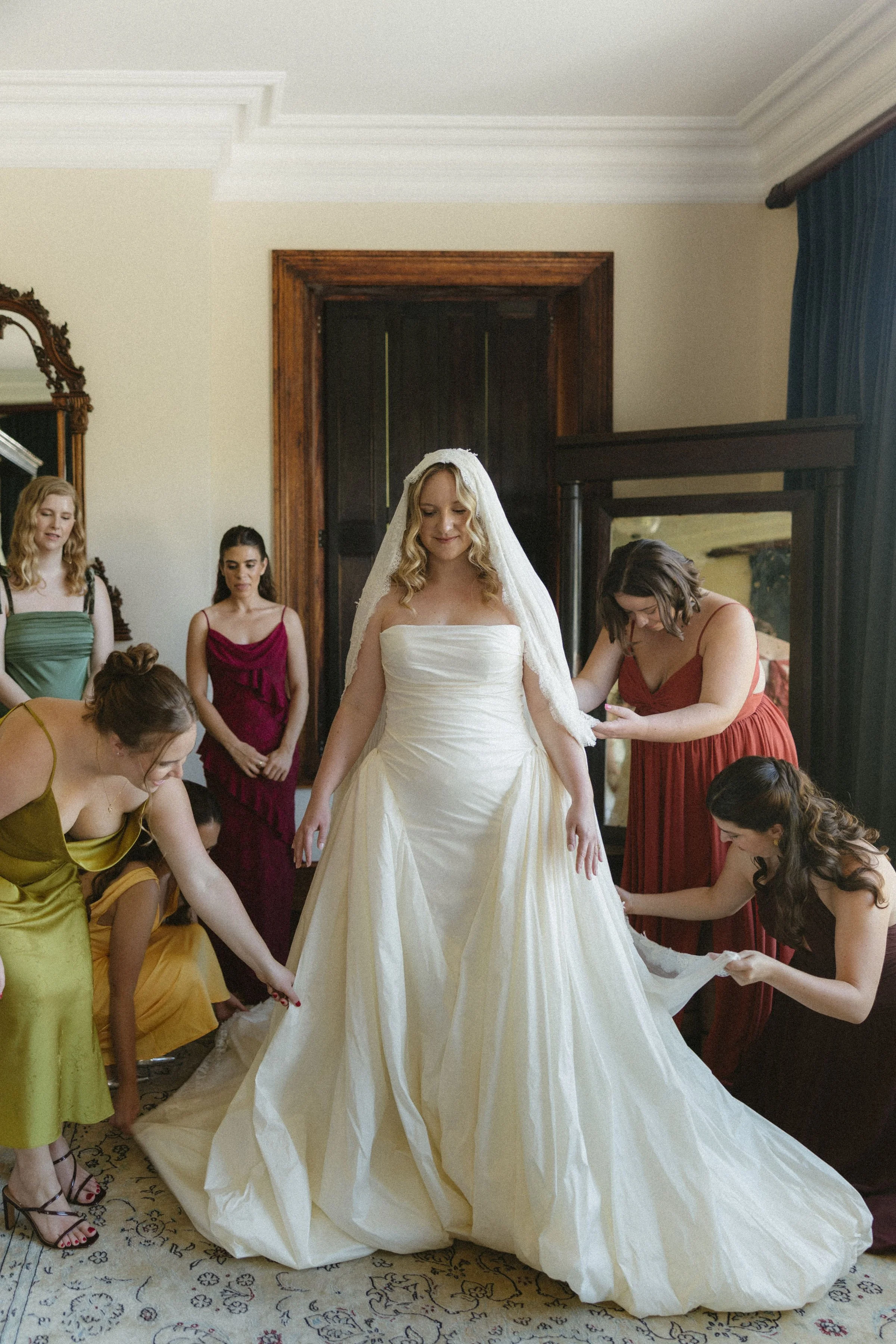 Bride in a white wedding gown and veil surrounded by bridesmaids adjusting her dress indoors