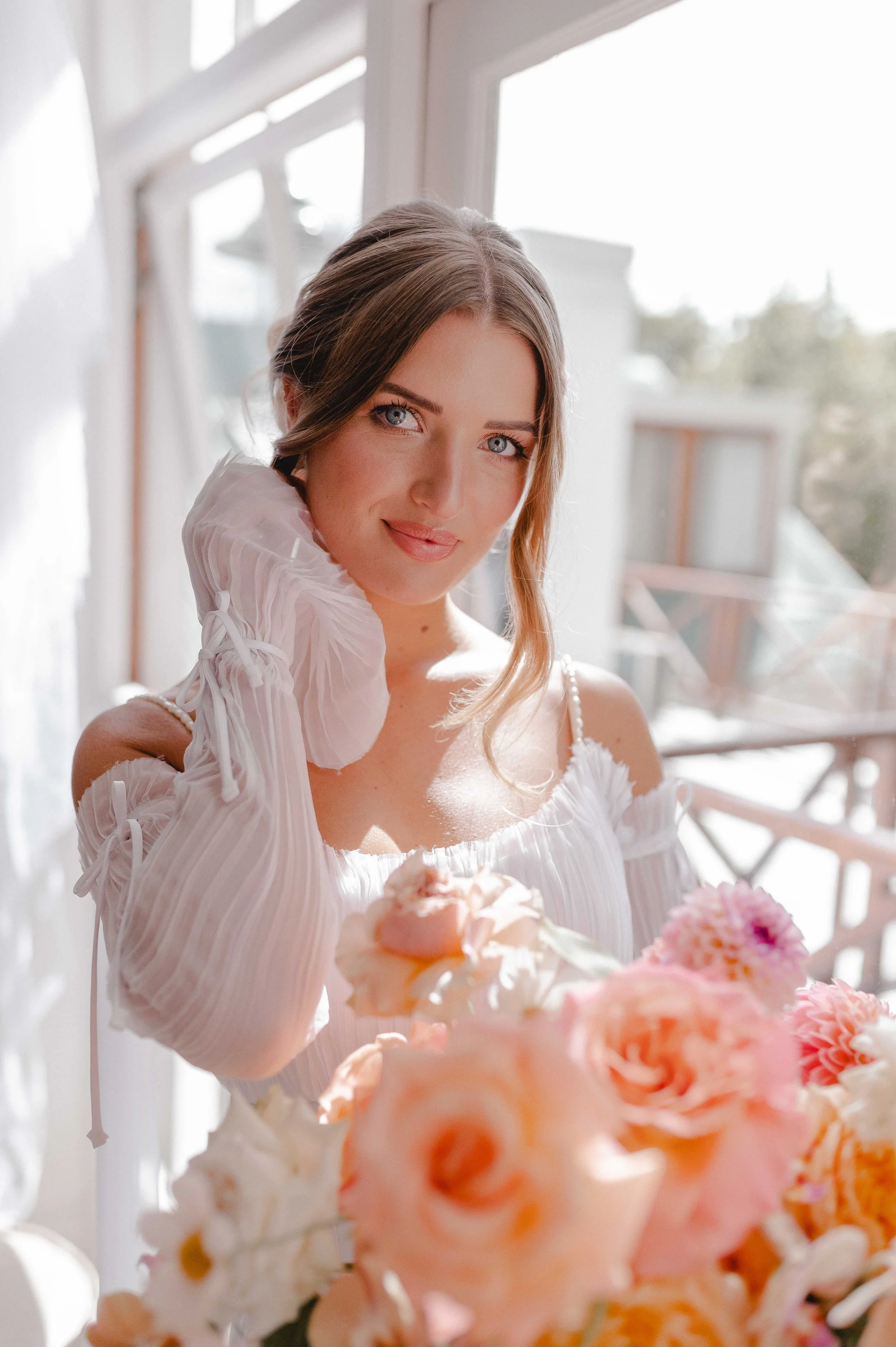 Smiling woman in a white dress holding a pastel flower bouquet in bright window light