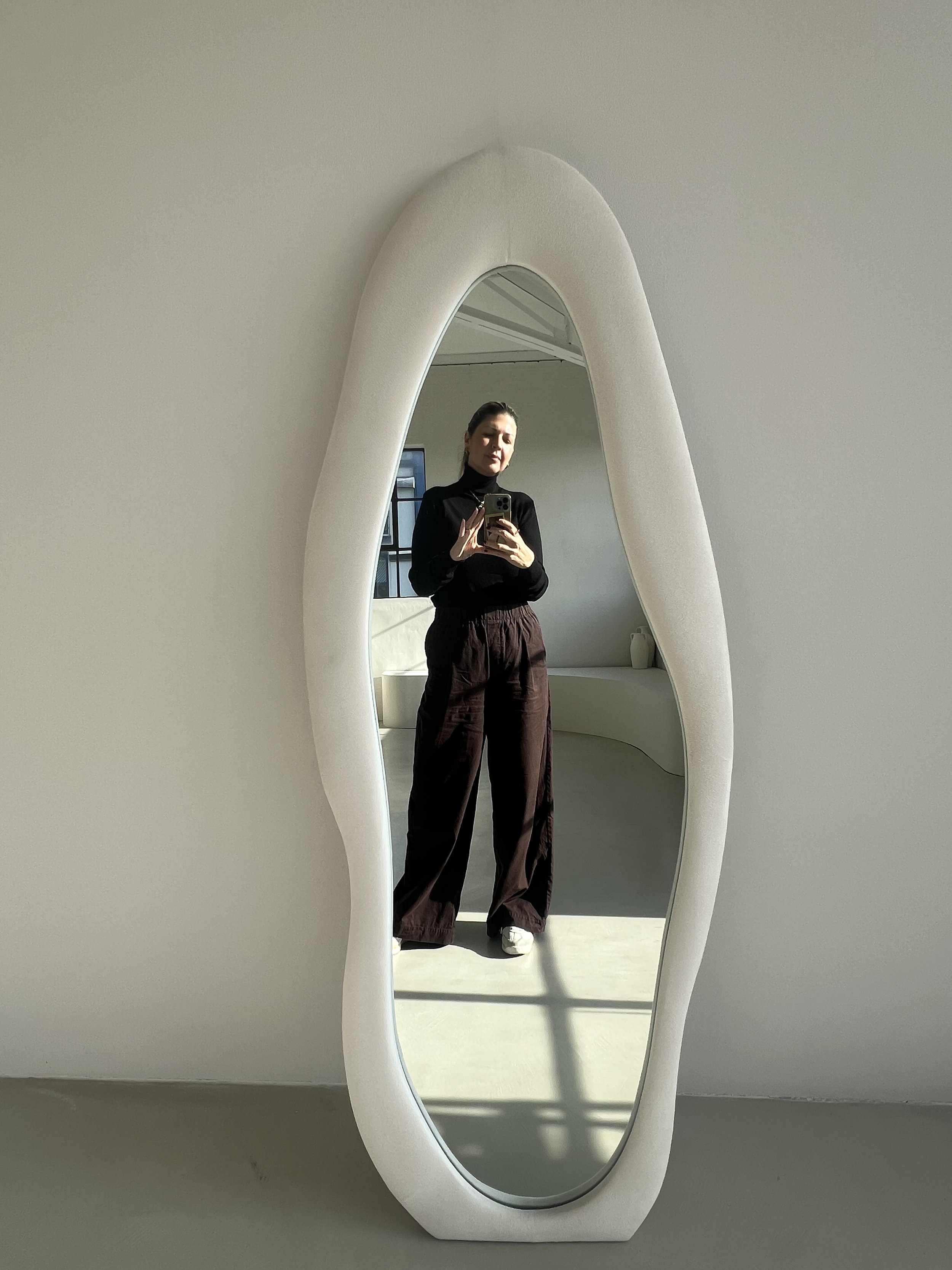 Woman taking a full-length mirror selfie in a bright modern interior, wearing a black turtleneck and loose brown pants.