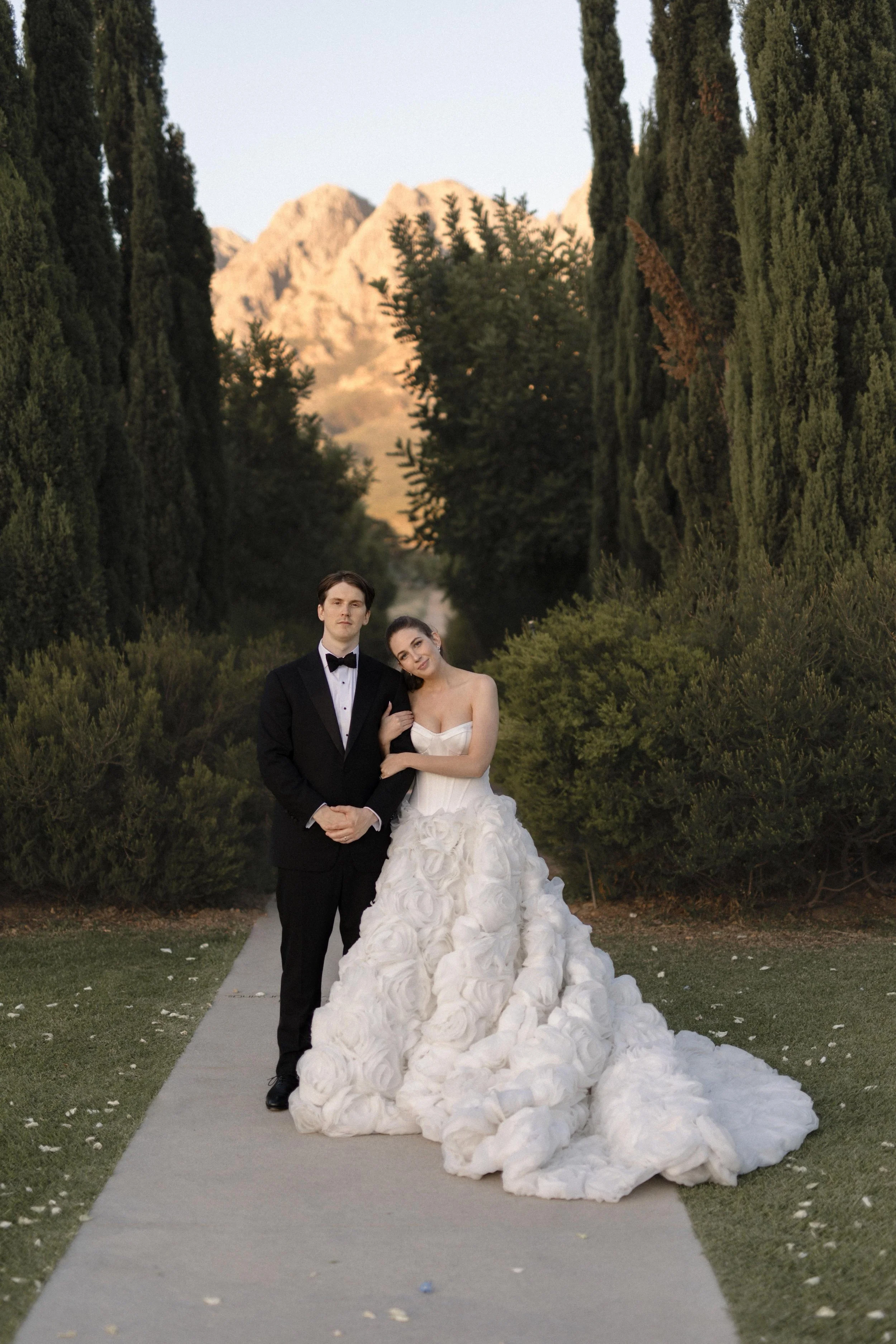 Bride and groom posing on a garden walkway with tall cypress trees and sunlit mountains in the background