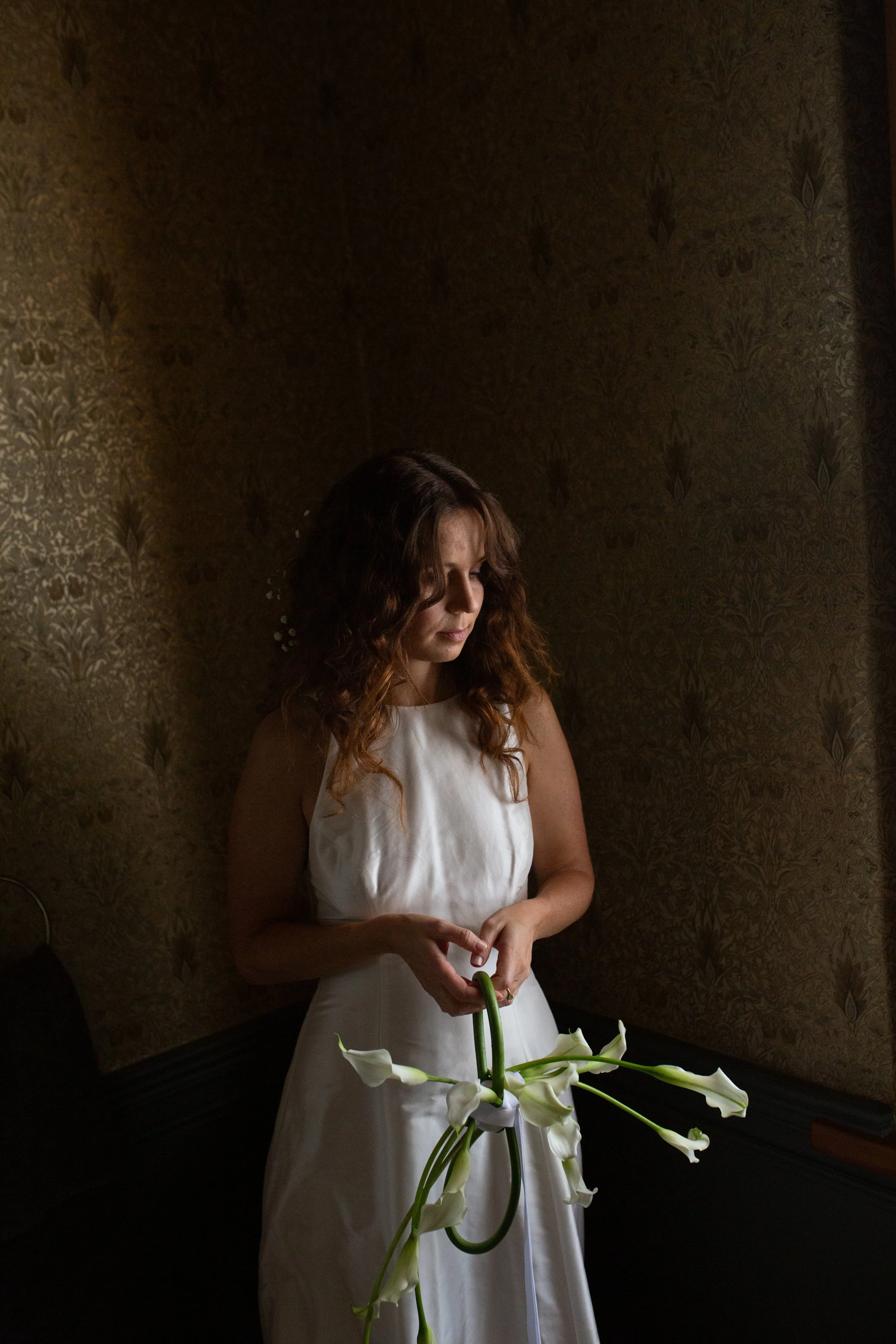 Woman in a white dress holding white calla lilies against dark vintage floral wallpaper