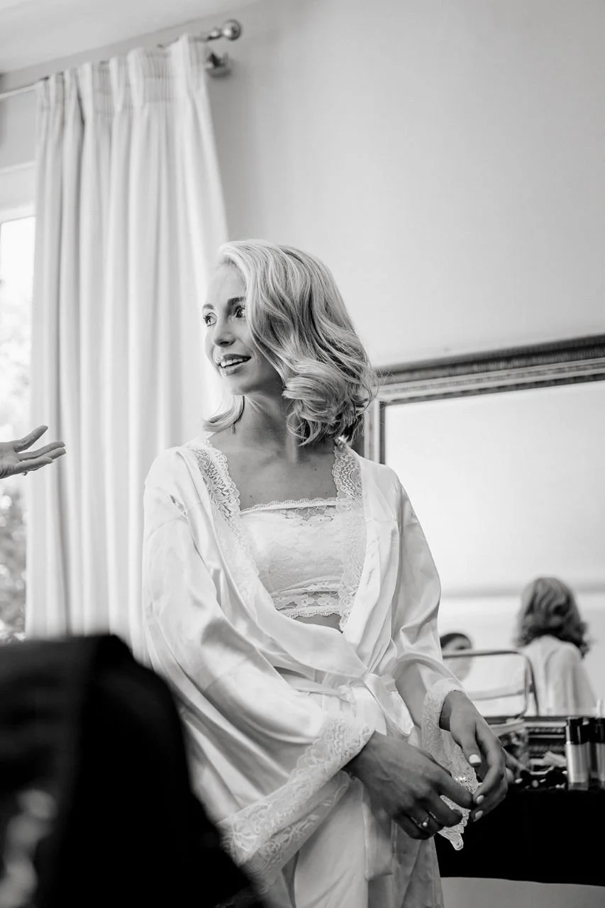 Black-and-white portrait of a smiling blonde woman in a lace-trimmed silk robe sitting in a bright bedroom.