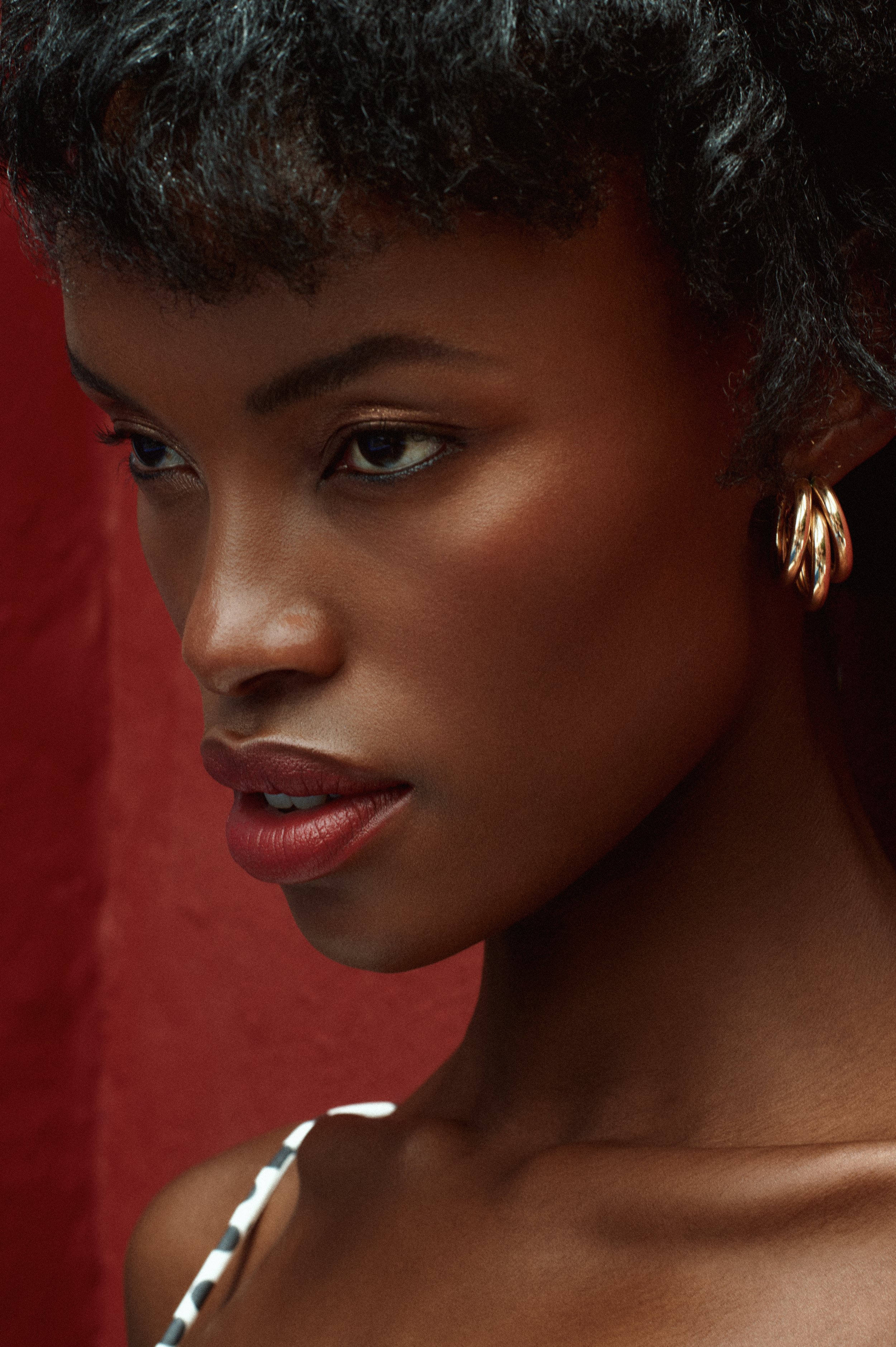 Close-up portrait of a Black woman with natural curly hair, gold hoop earrings, and red lipstick against a red background