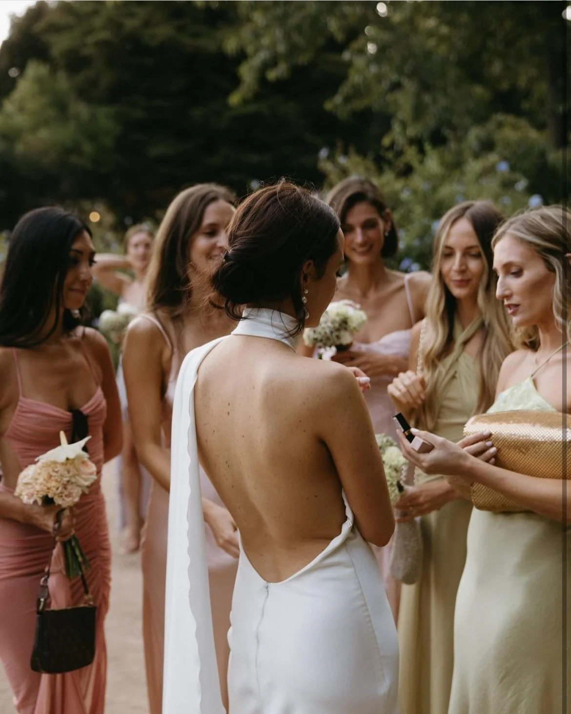 Bride in a backless white wedding dress surrounded by bridesmaids holding bouquets at an outdoor wedding