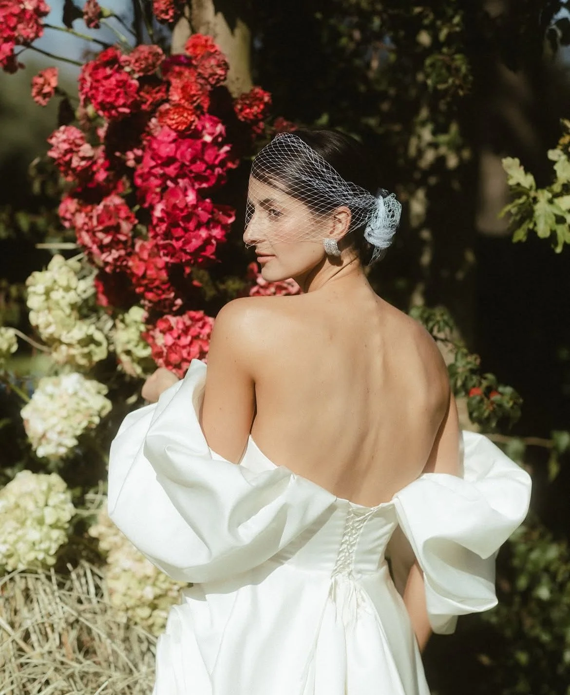 Bride in a white off-the-shoulder wedding dress and birdcage veil posing in a flower garden