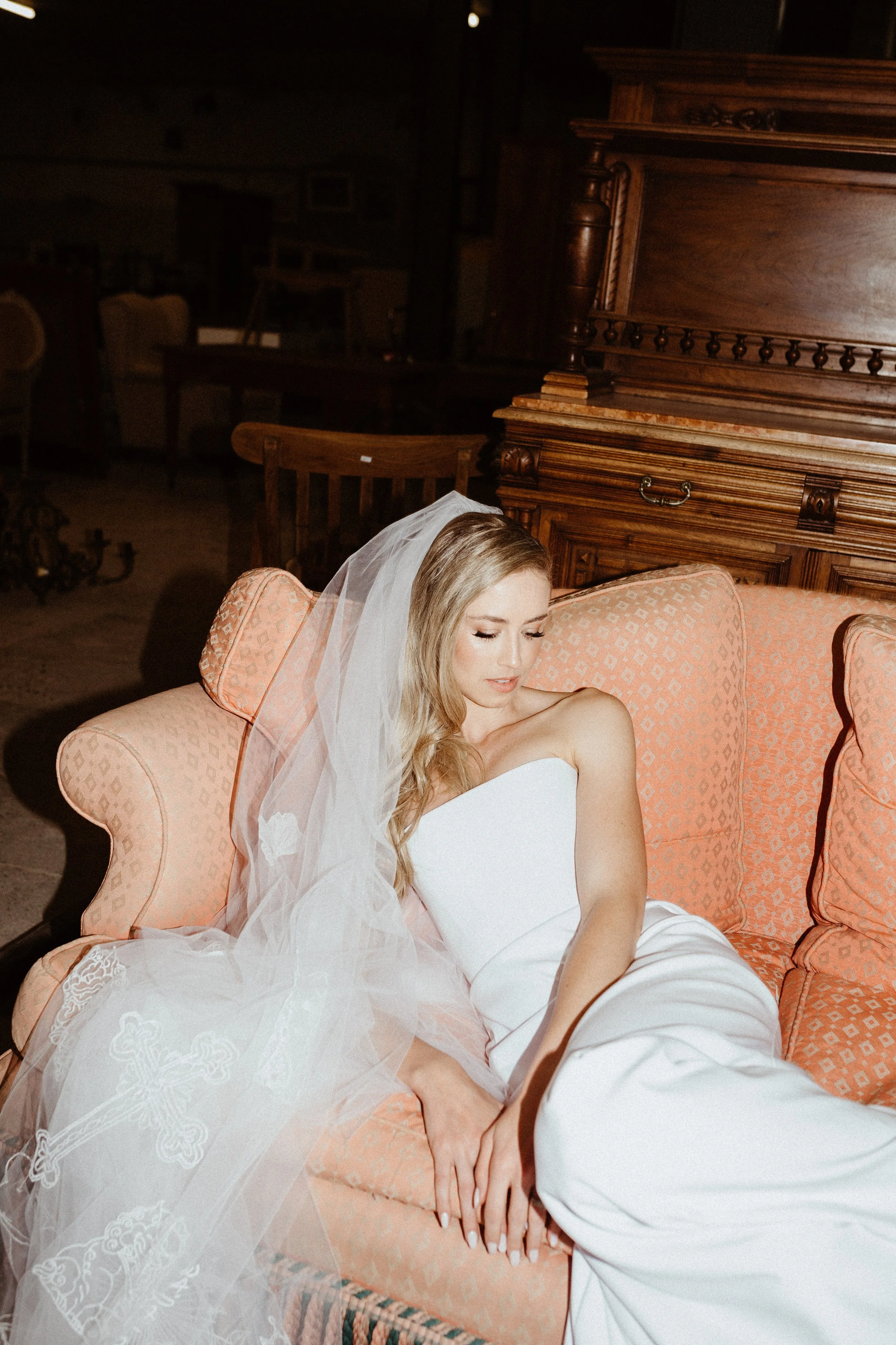 Bride in a white wedding dress and veil sitting on a vintage pink sofa beside an ornate wooden antique cabinet.