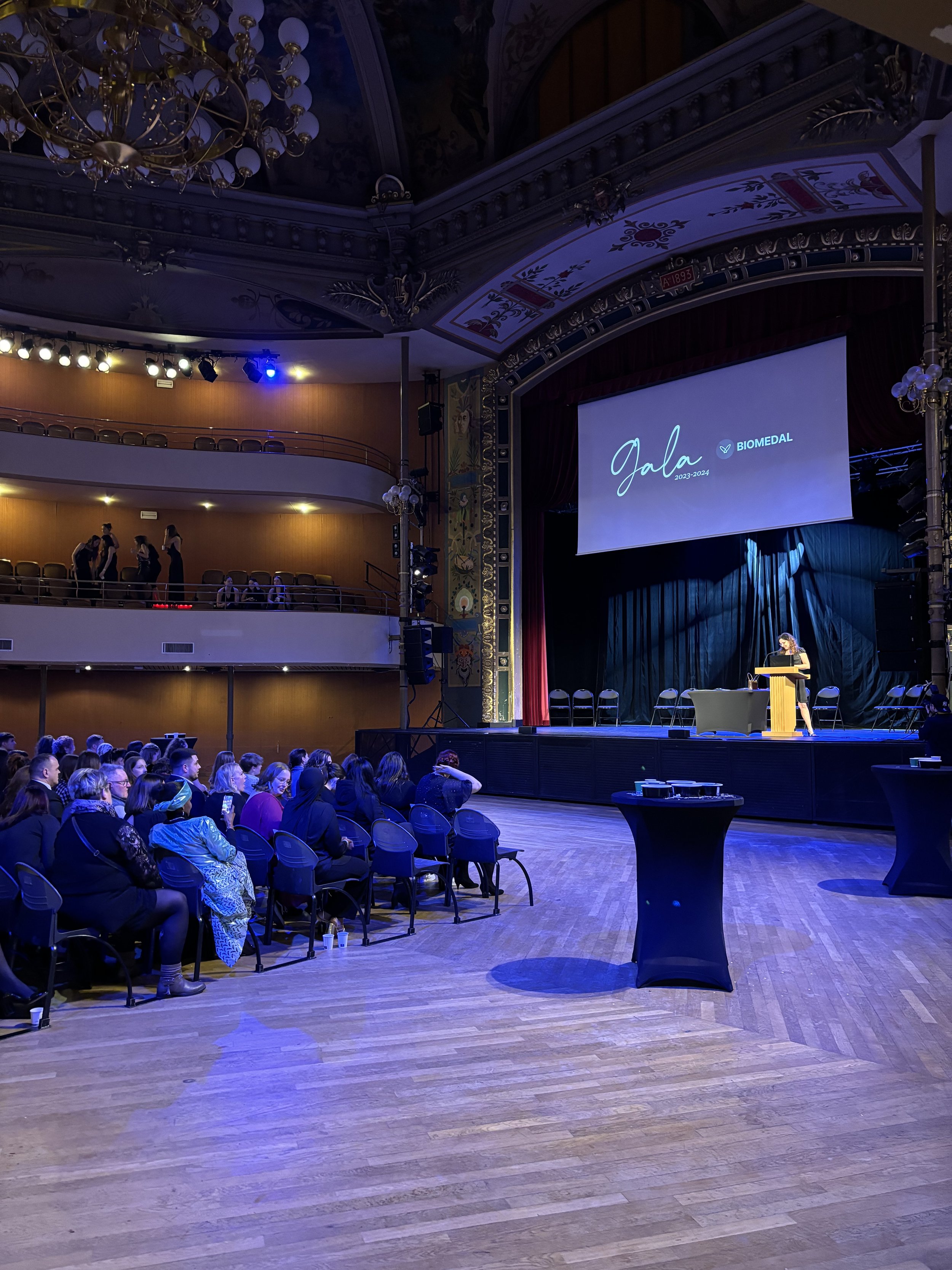 Cérémonie de gala dans un théâtre historique, avec une intervenante sur scène, un public assis et un écran affichant « Gala Biomedal 2023-2024 ».